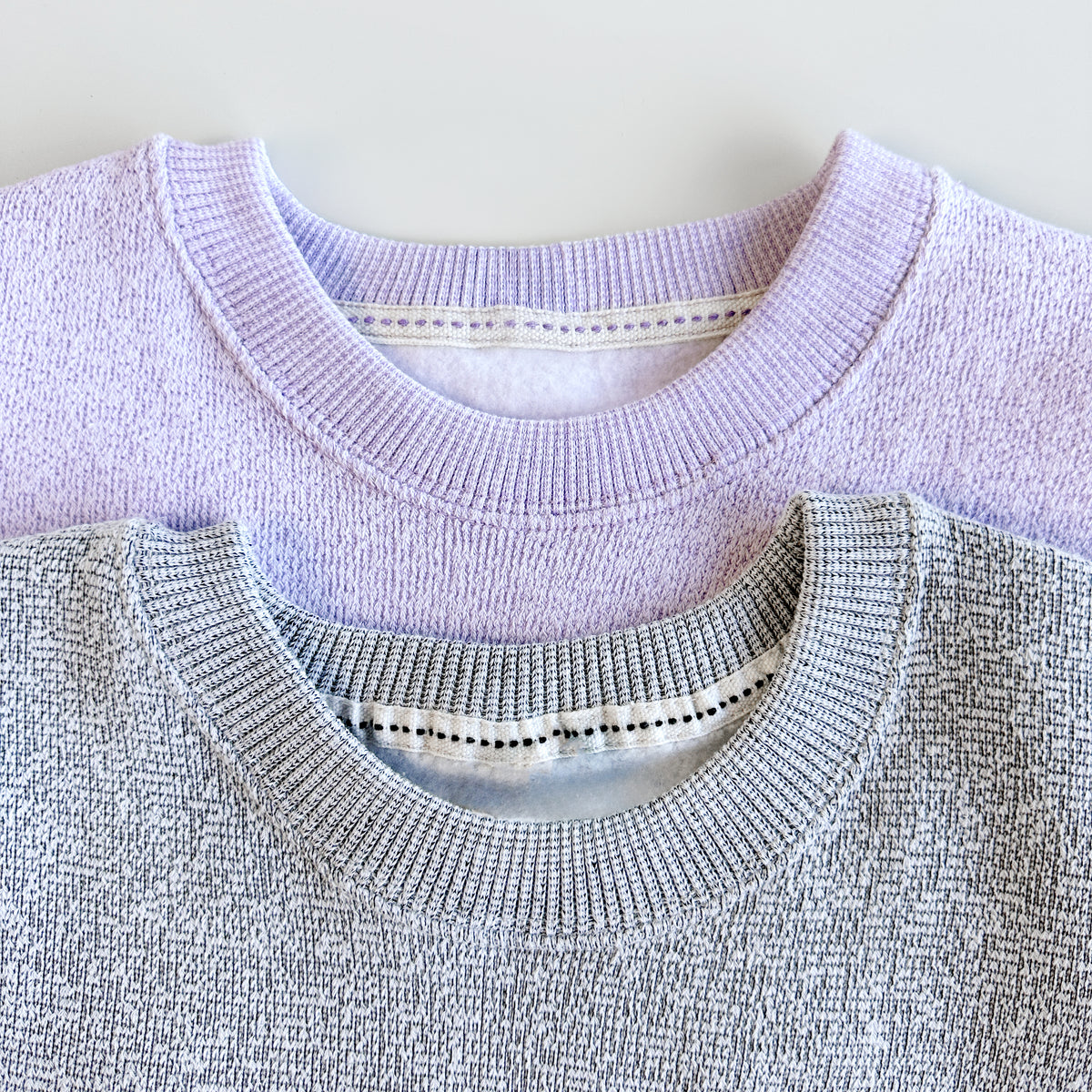 Lavender and heather grey knit sweatshirts stacked together, showing their ribbed crew necklines.