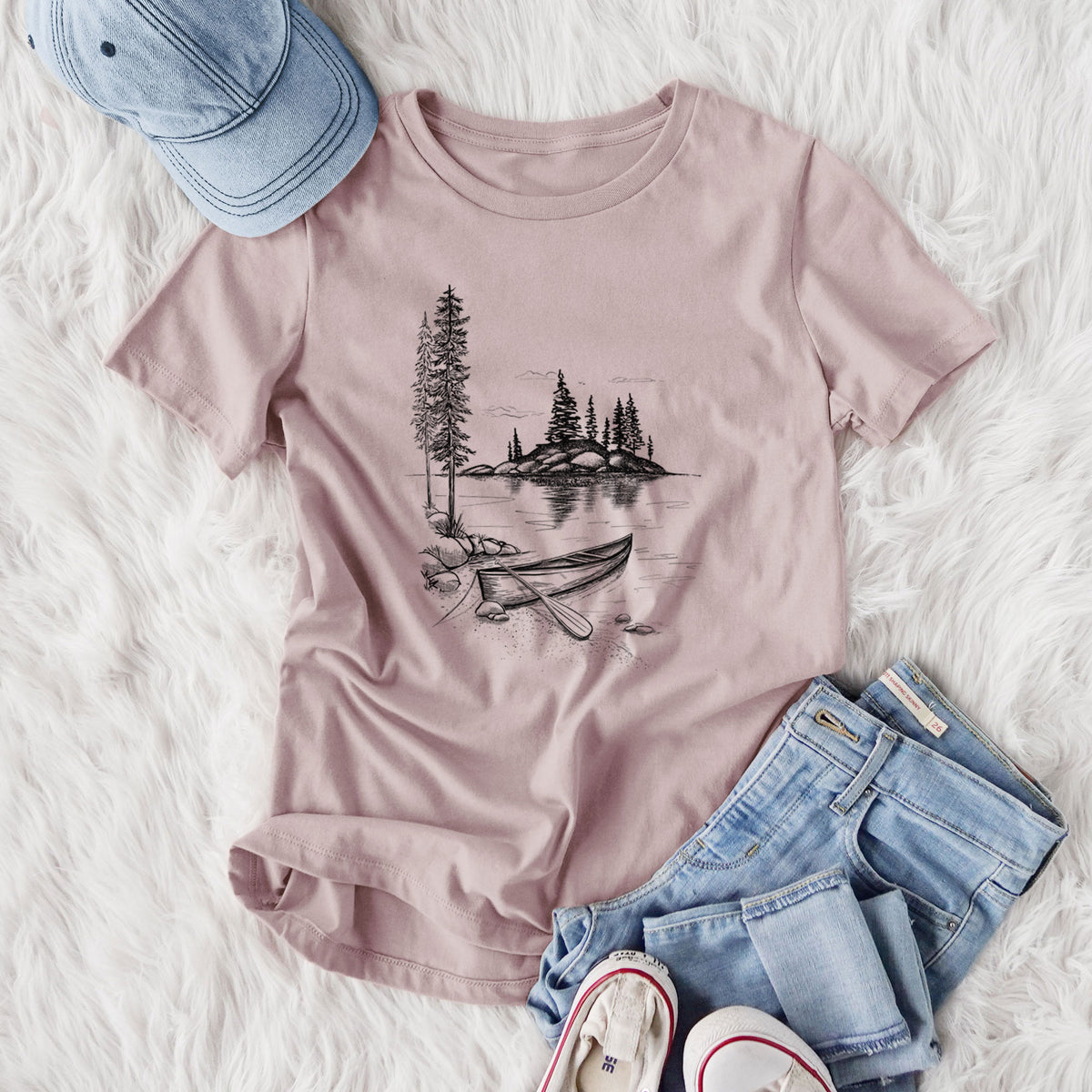 Women's relaxed fit cotton t-shirt printed with a black ink illustration of a canoe resting on the shore of a lake with pine trees on a small island, styled flat on a white faux fur rug with jeans.