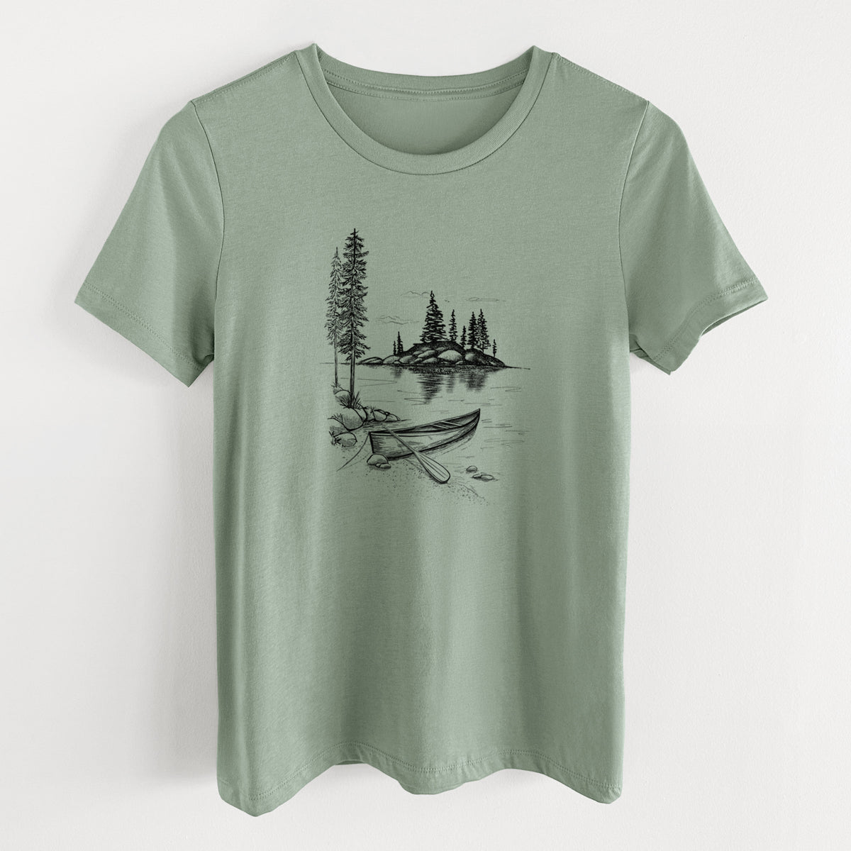 Sage green women's relaxed fit cotton t-shirt laid flat, printed with a black ink illustration of a canoe resting on the shore of a lake with pine trees on a small island.