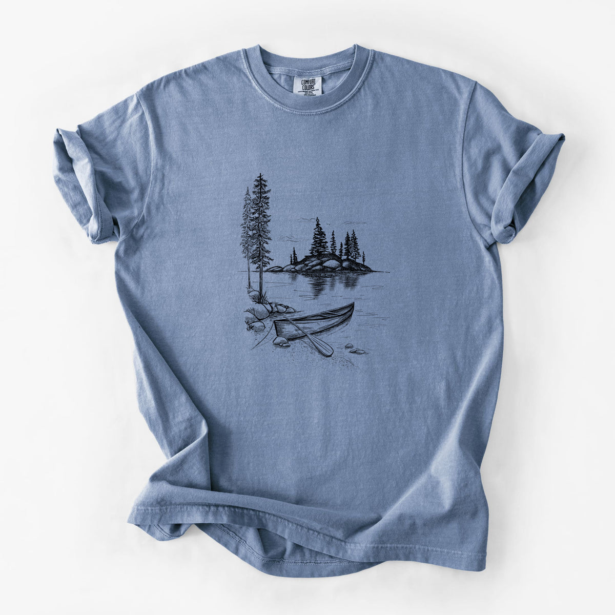 Blue jean Comfort Colors t-shirt laid flat, printed with a black ink illustration of a canoe resting on the shore of a lake with pine trees on a small island.