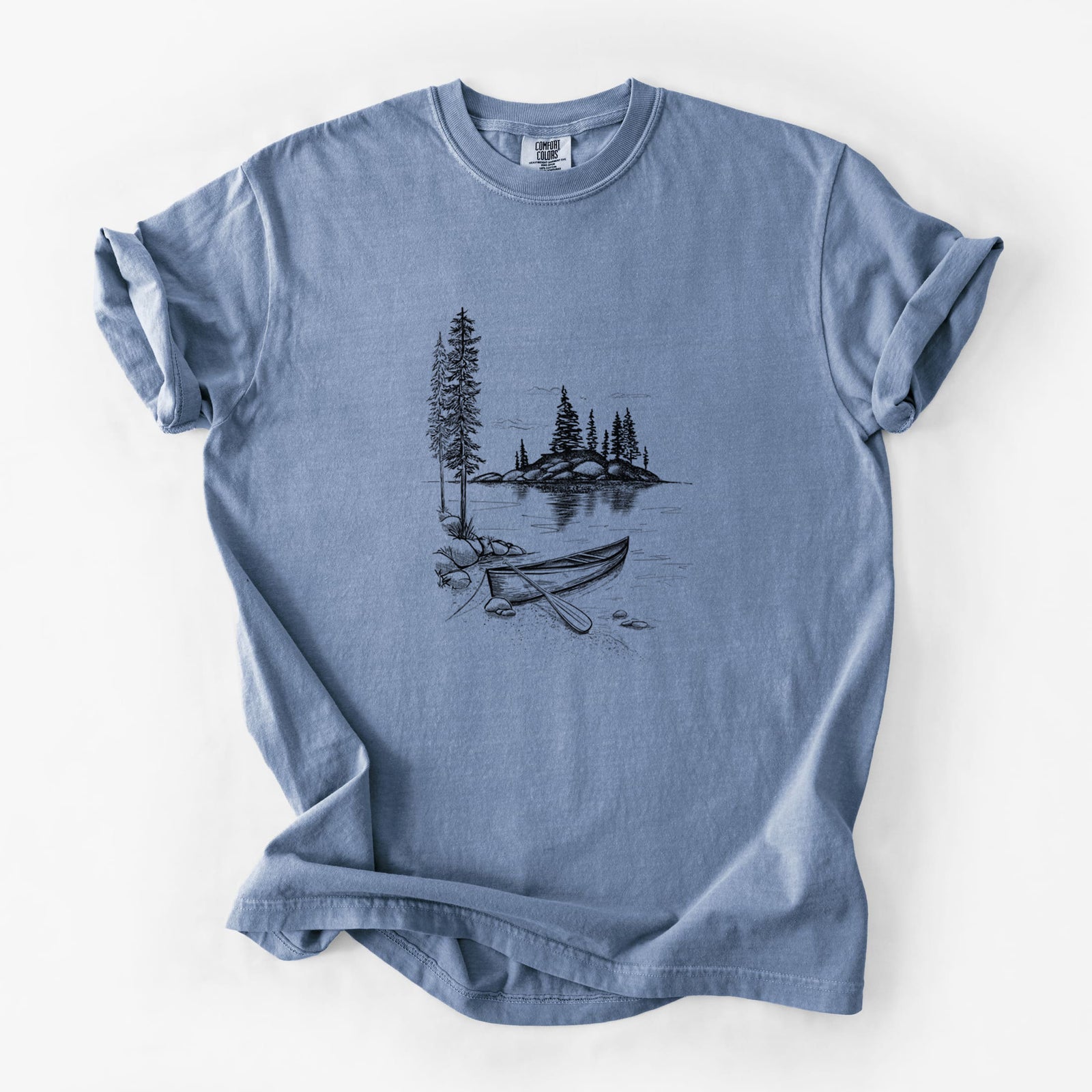 Blue jean Comfort Colors t-shirt laid flat, printed with a black ink illustration of a canoe resting on the shore of a lake with pine trees on a small island.
