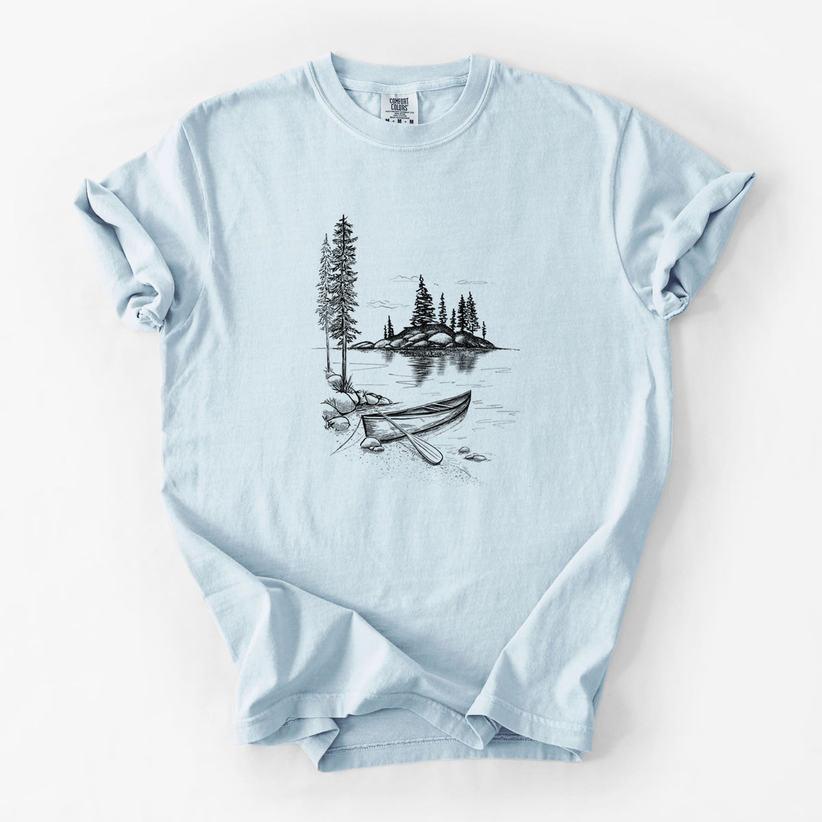 Chambray blue Comfort Colors t-shirt laid flat, printed with a black ink illustration of a canoe resting on the shore of a lake with pine trees on a small island.