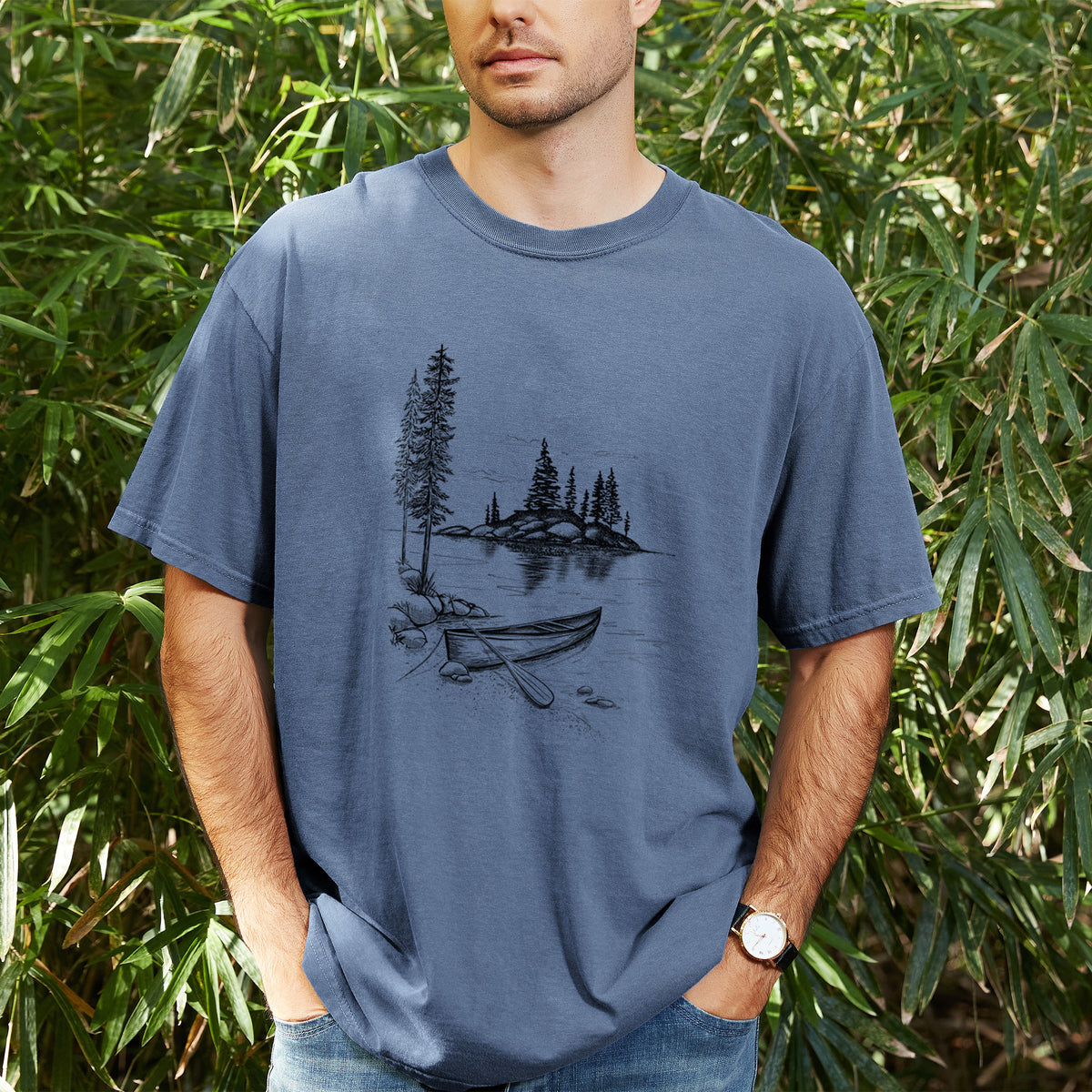 Man wearing a Comfort Colors t-shirt printed with a black ink illustration of a canoe resting on the shore of a lake with pine trees on a small island, outdoors.