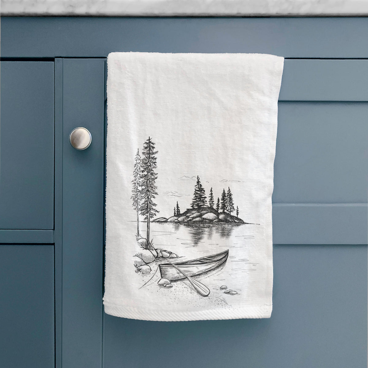 White hand towel printed with a black ink illustration of a canoe resting on the shore of a lake with pine trees on a small island, hanging on a cabinet door.