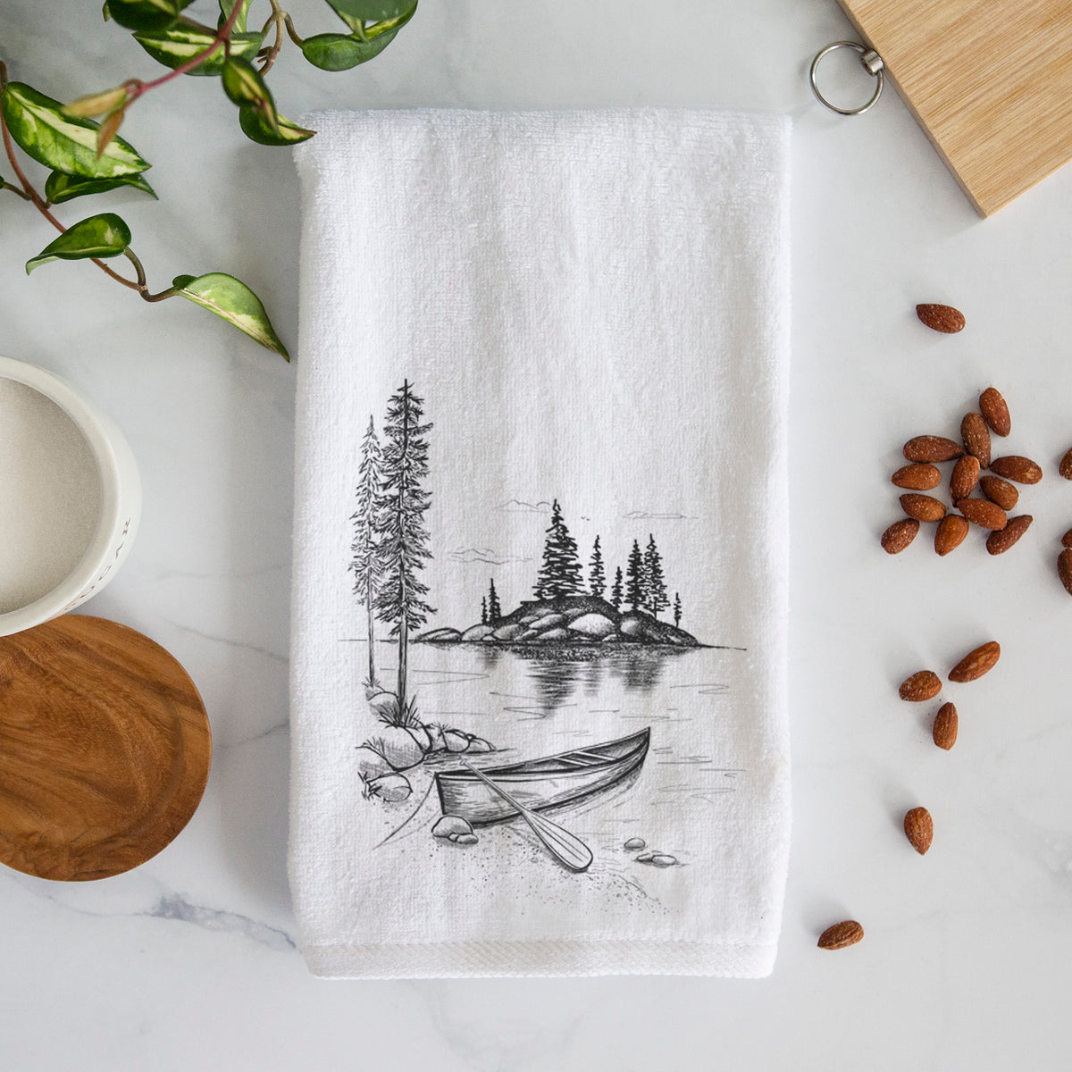 White hand towel printed with a black ink illustration of a canoe resting on the shore of a lake with pine trees on a small island.