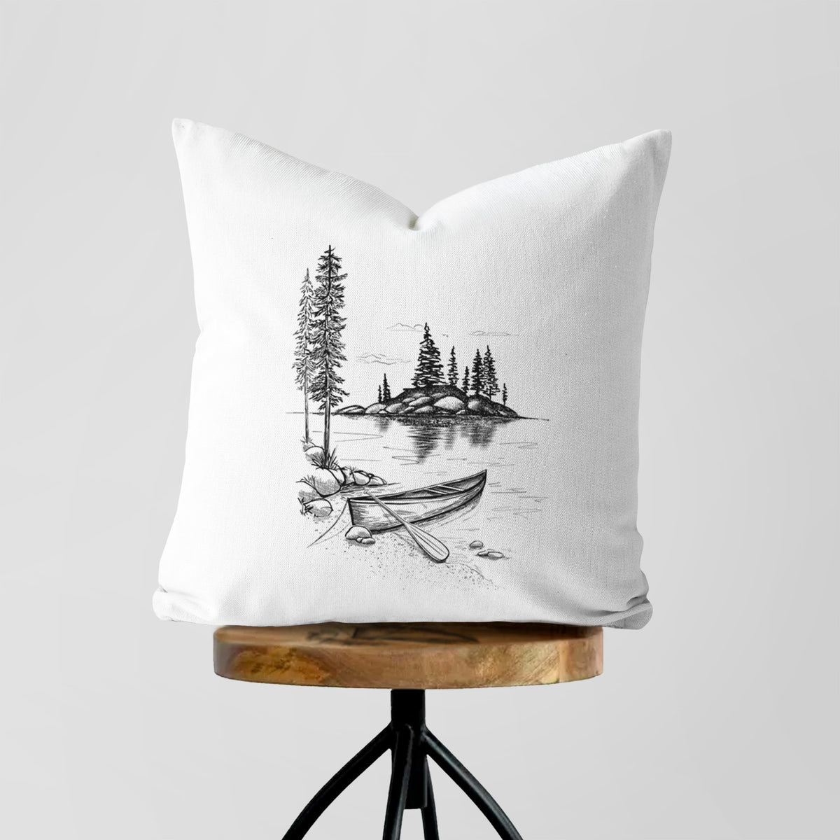 White pillow cover printed with a black ink illustration of a canoe resting on the shore of a lake with pine trees on a small island, on a wooden stool.