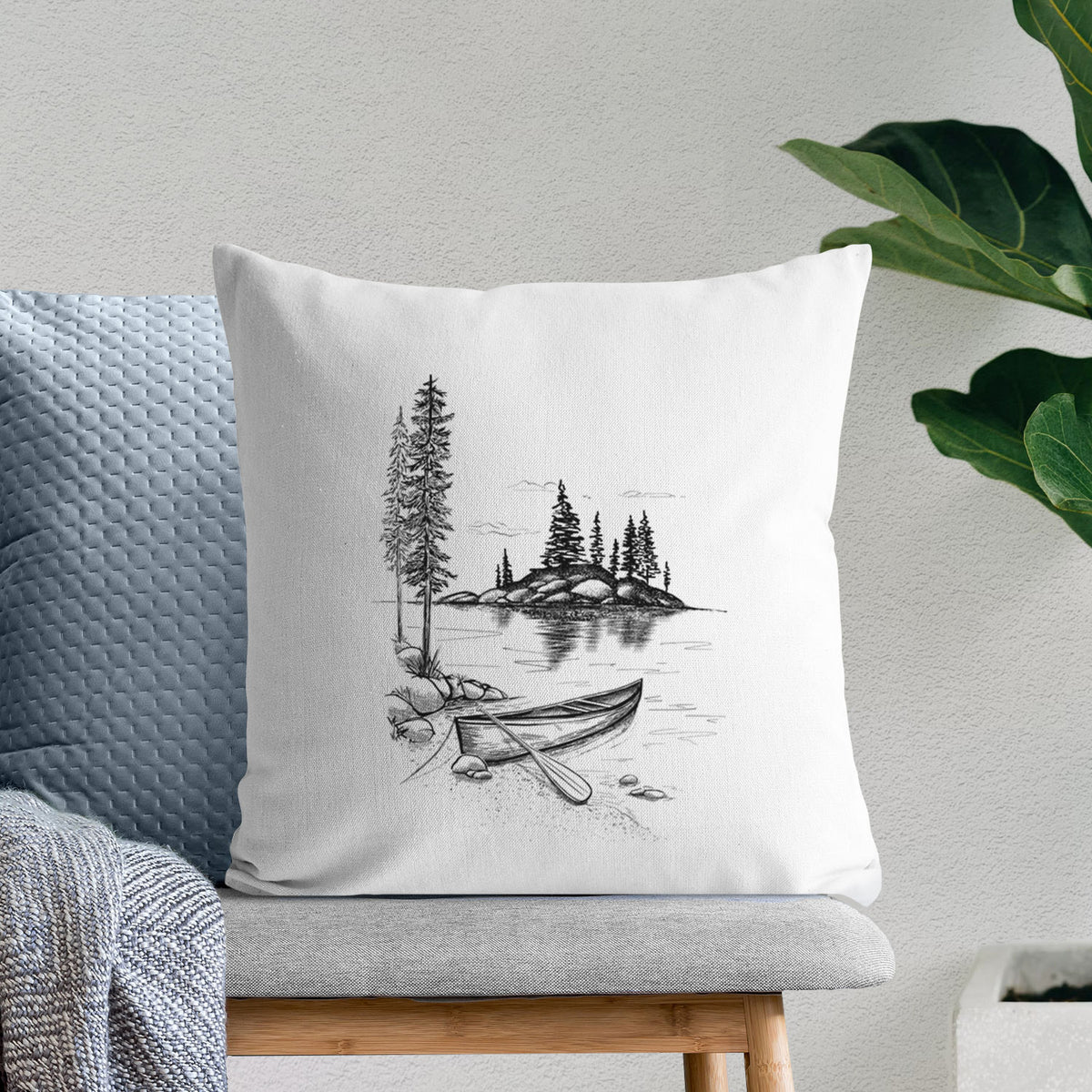 White pillow cover printed with a black ink illustration of a canoe resting on the shore of a lake with pine trees on a small island, on a bench beside plants.