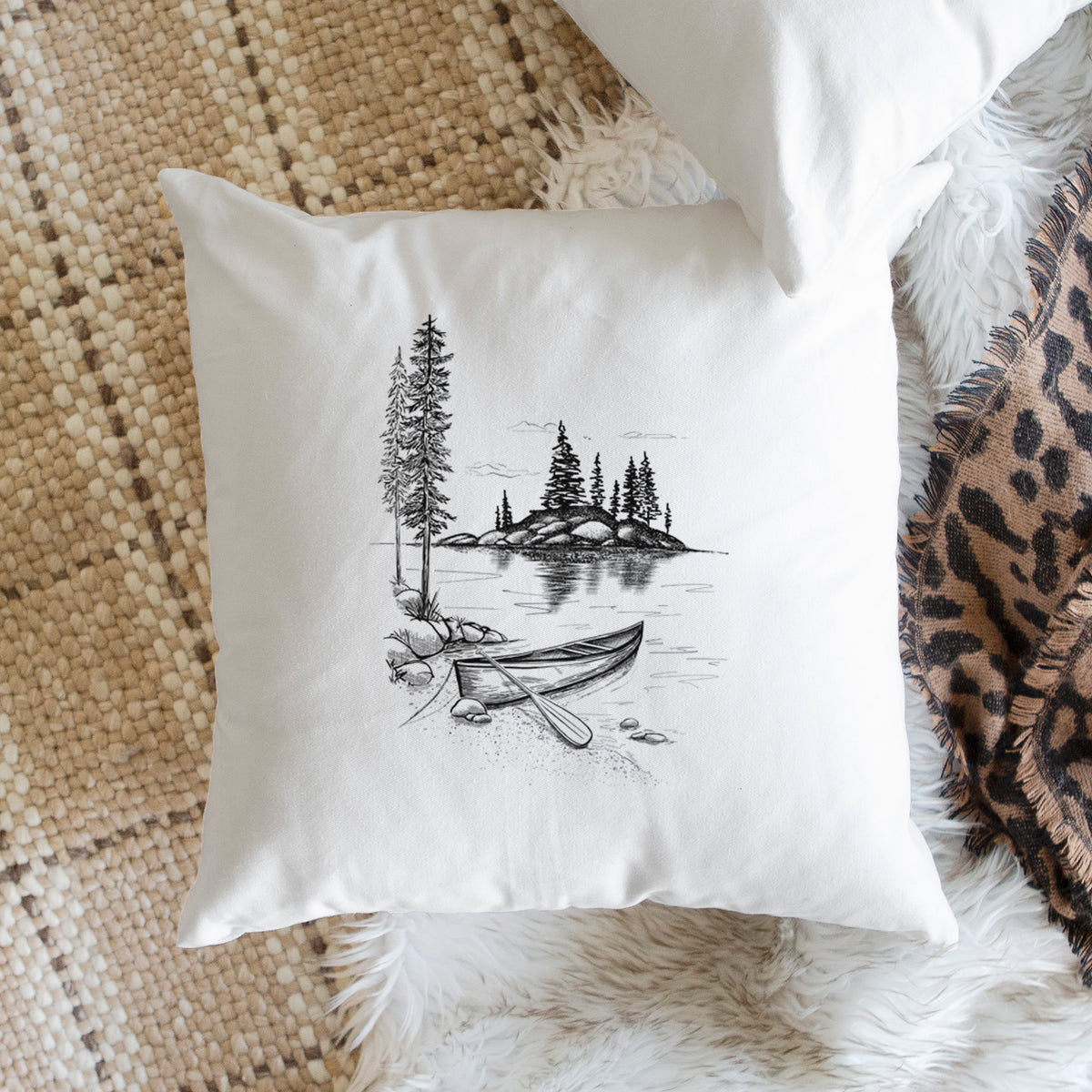 White pillow cover printed with a black ink illustration of a canoe resting on the shore of a lake with pine trees on a small island, on a woven chair.