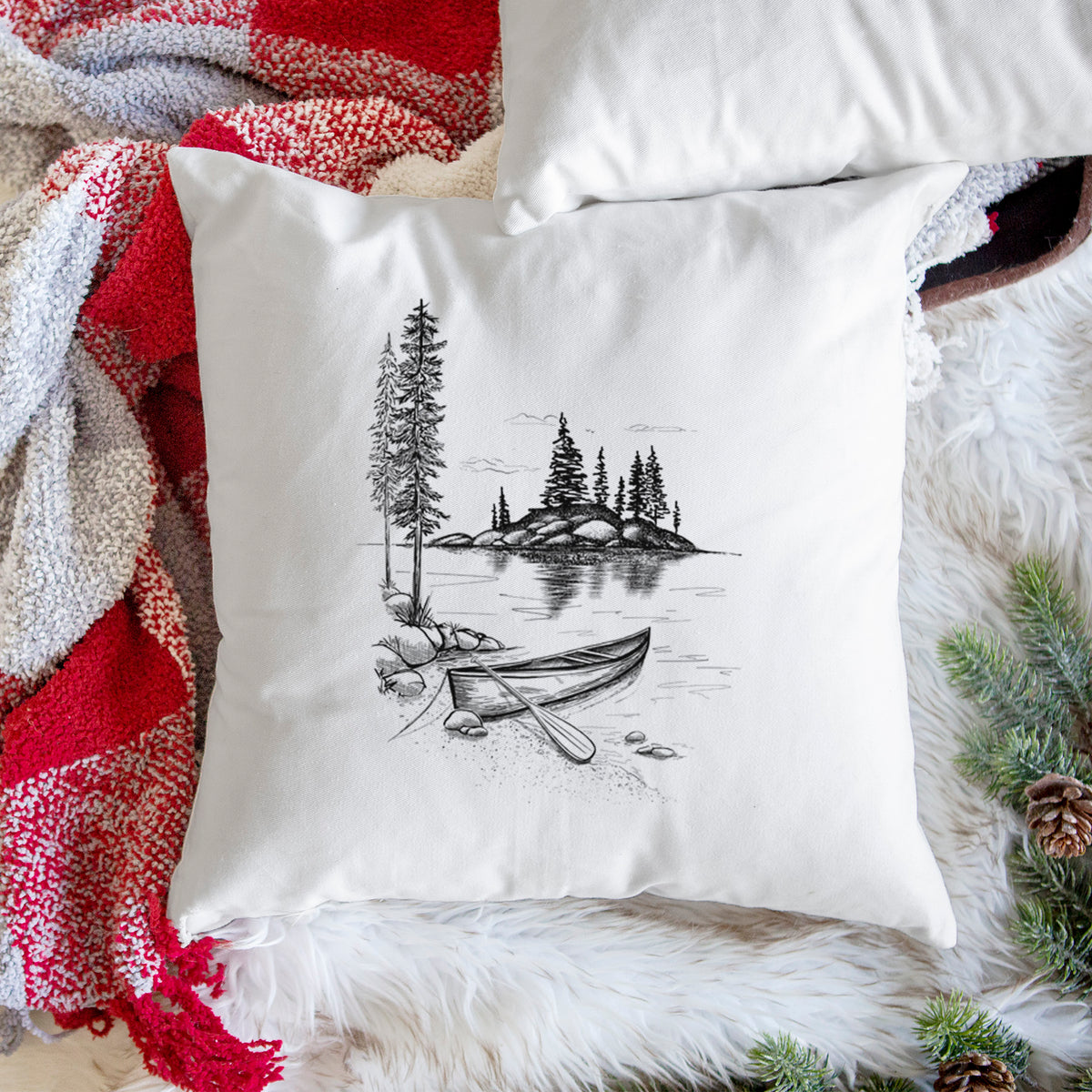 White pillow cover printed with a black ink illustration of a canoe resting on the shore of a lake with pine trees on a small island, on a woven chair.
