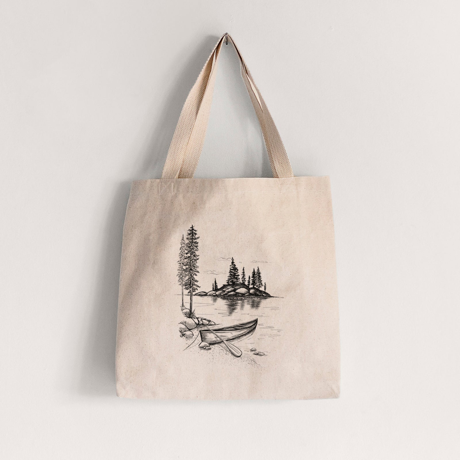 Natural canvas tote bag printed with a black ink illustration of a canoe resting on the shore of a lake with pine trees on a small island.