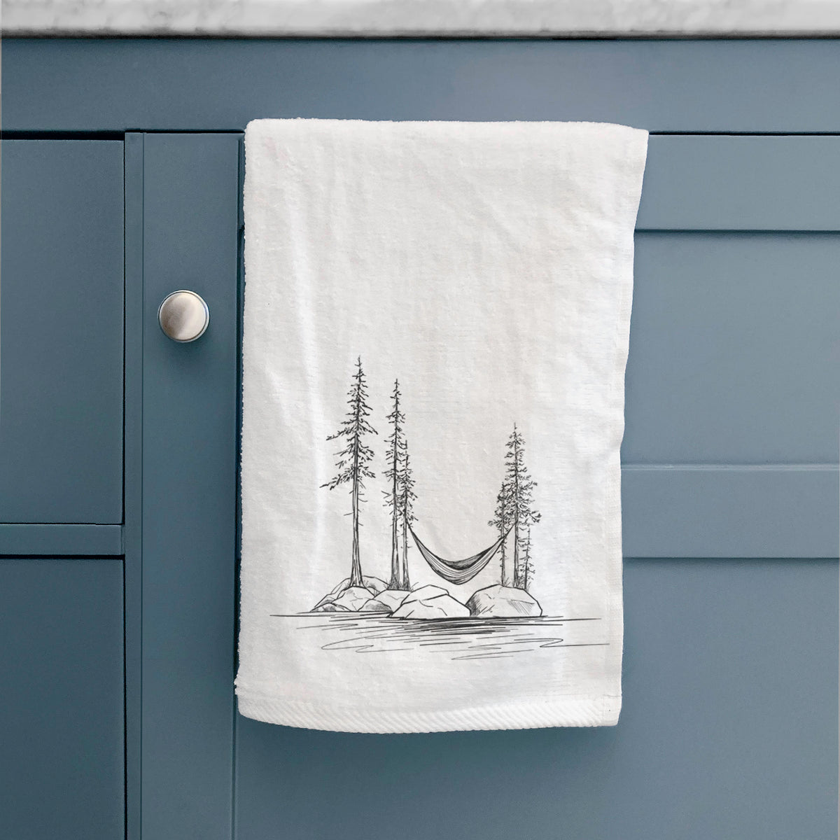 White hand towel printed with a black ink illustration of a hammock strung between pine trees on a small lakeside island, hanging on a cabinet door.