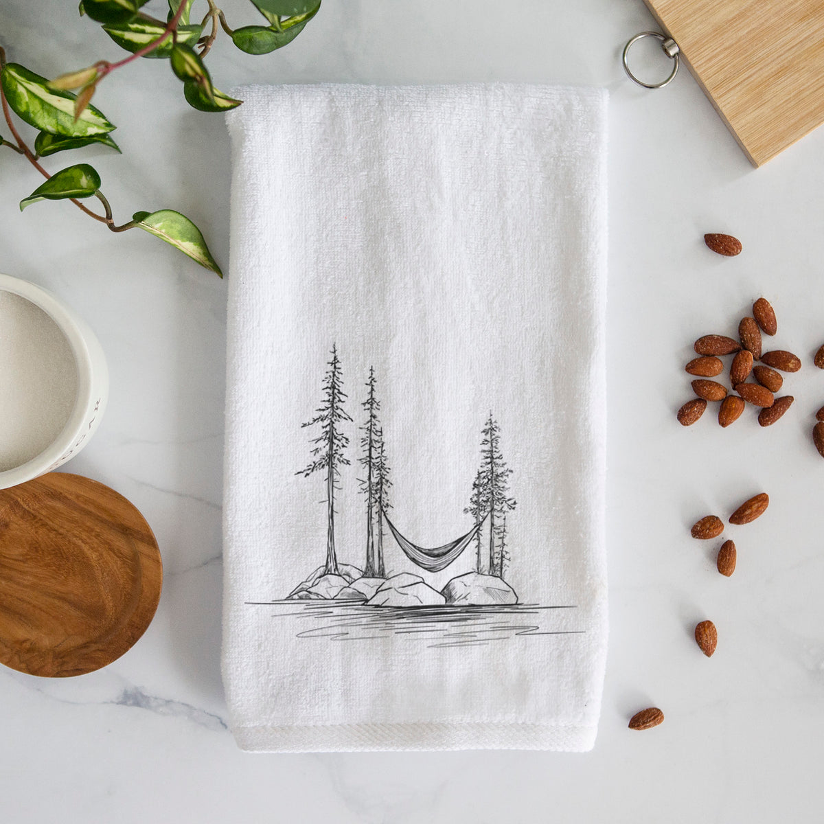 White hand towel printed with a black ink illustration of a hammock strung between pine trees on a small lakeside island.