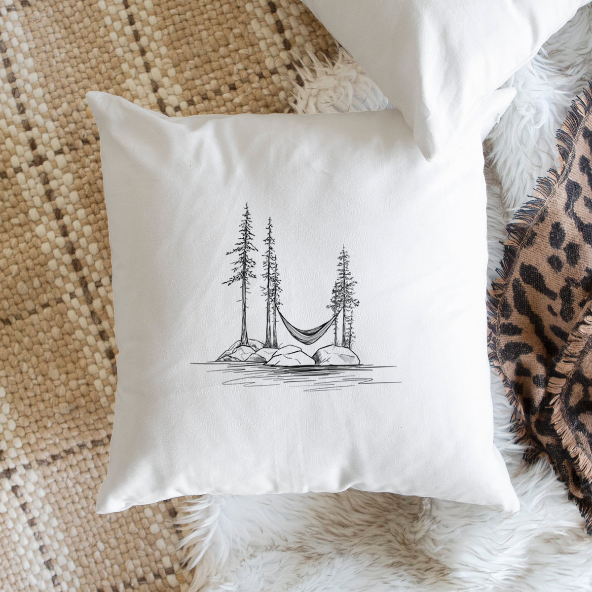White pillow cover printed with a black ink illustration of a hammock strung between pine trees on a small lakeside island, on a woven chair.
