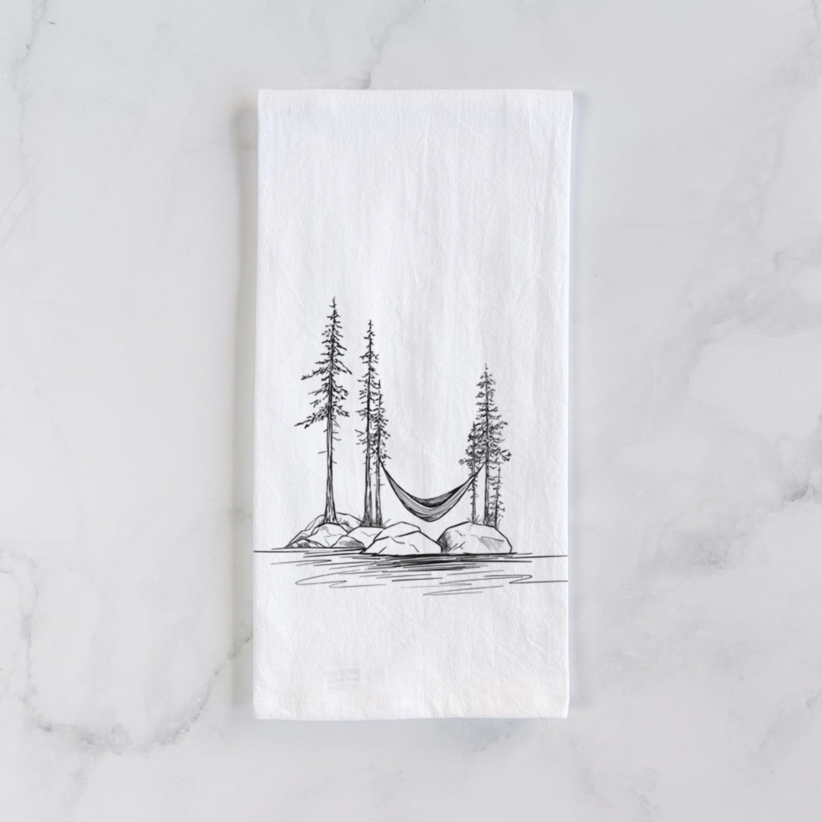 White cotton tea towel laid flat on marble, printed with a black ink illustration of a hammock strung between pine trees on a small lakeside island.