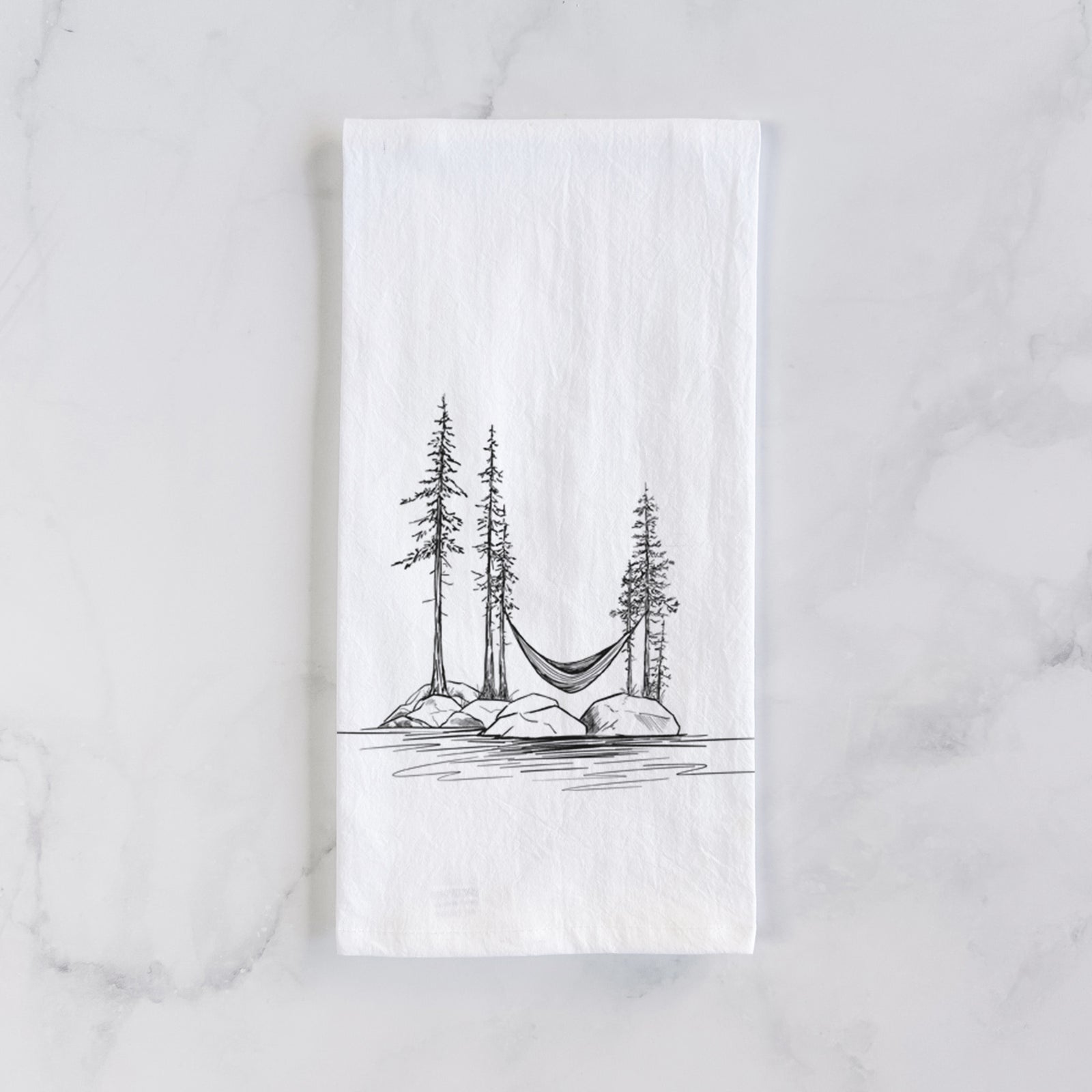 White cotton tea towel printed with a black ink illustration of a hammock strung between pine trees on a small lakeside island, on a marble counter beside a round wooden cutting board and greenery.