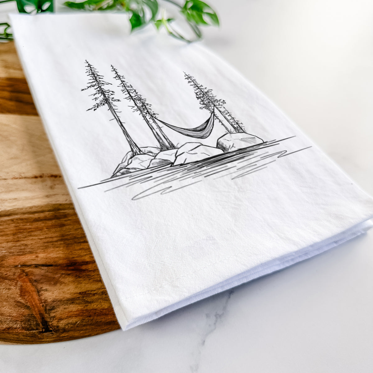 Close-up of a folded white tea towel on a wooden board, showing a black ink illustration of a hammock strung between pine trees on a small lakeside island.