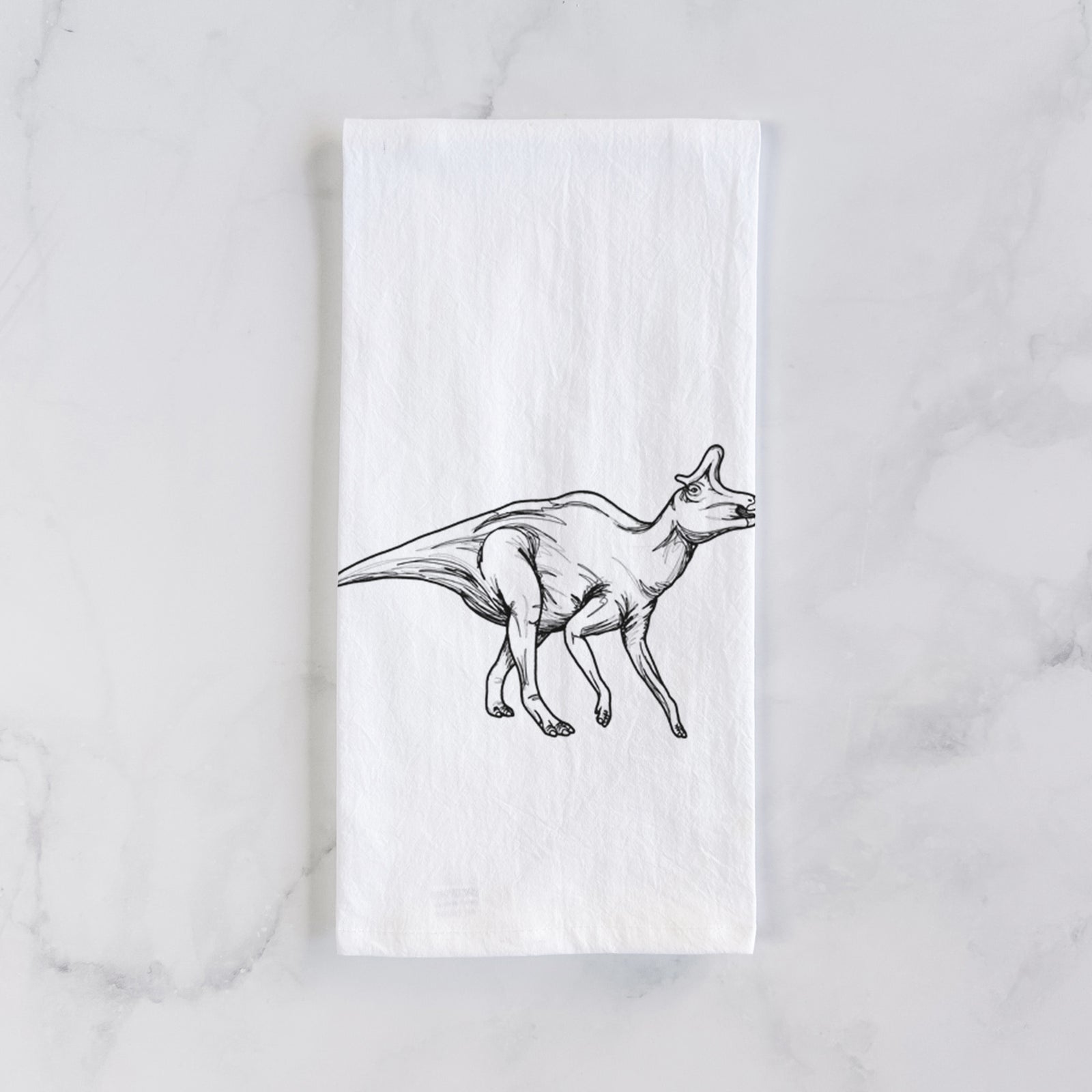 White cotton tea towel printed with a black ink illustration of a crested Lambeosaurus walking, on a marble counter beside a round wooden cutting board and greenery.