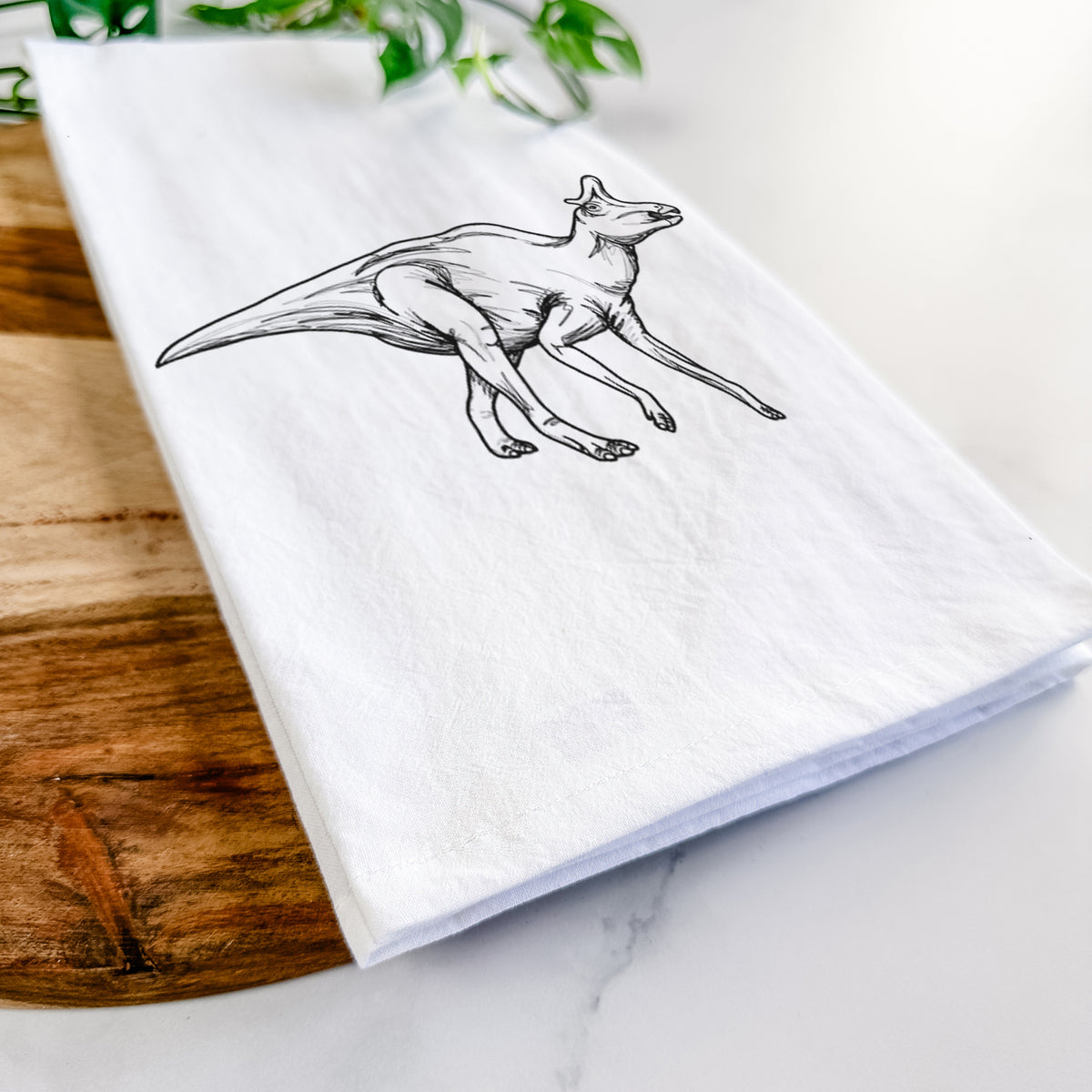 Close-up of a folded white tea towel on a wooden board, showing a black ink illustration of a crested Lambeosaurus walking.