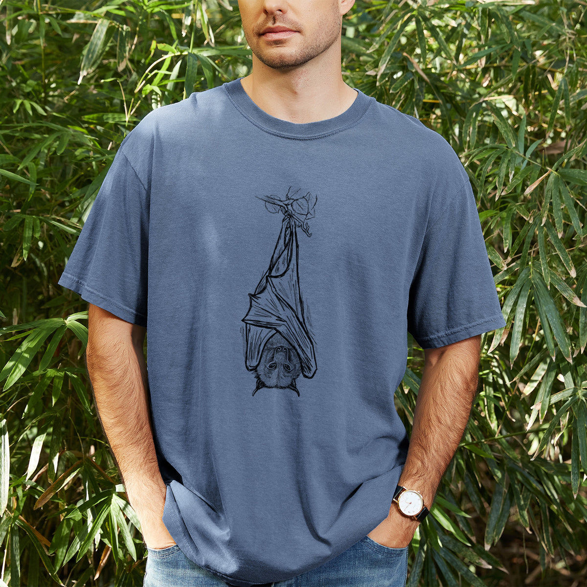 Man wearing a Comfort Colors t-shirt printed with a black ink illustration of a large flying fox bat hanging upside down with its wings wrapped around it, outdoors.