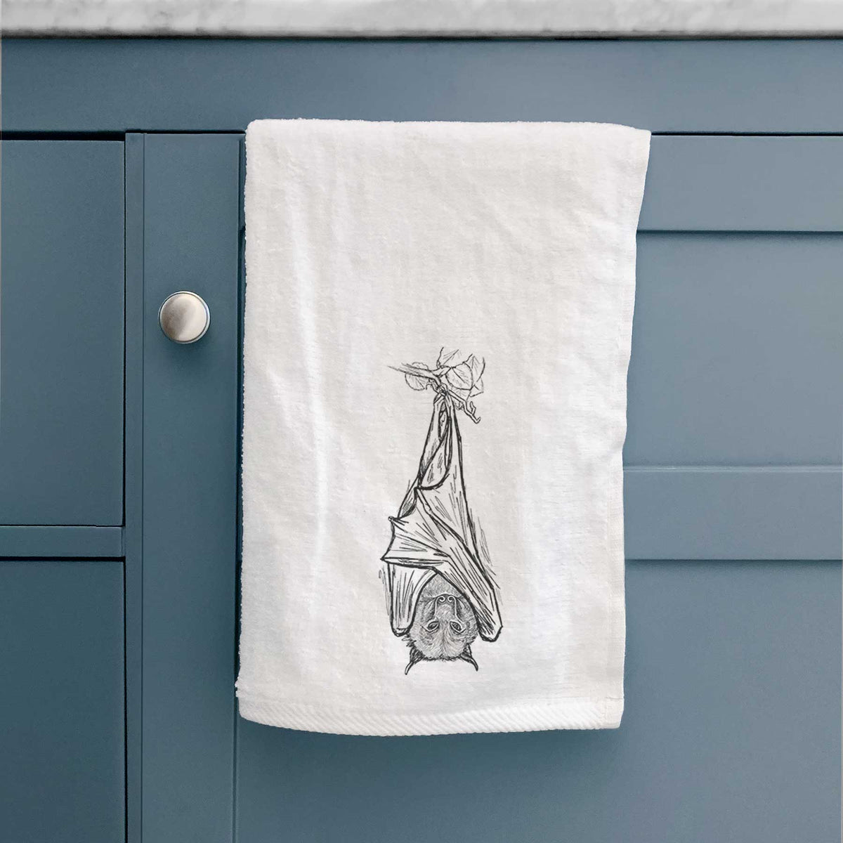 White hand towel printed with a black ink illustration of a large flying fox bat hanging upside down with its wings wrapped around it, hanging on a cabinet door.