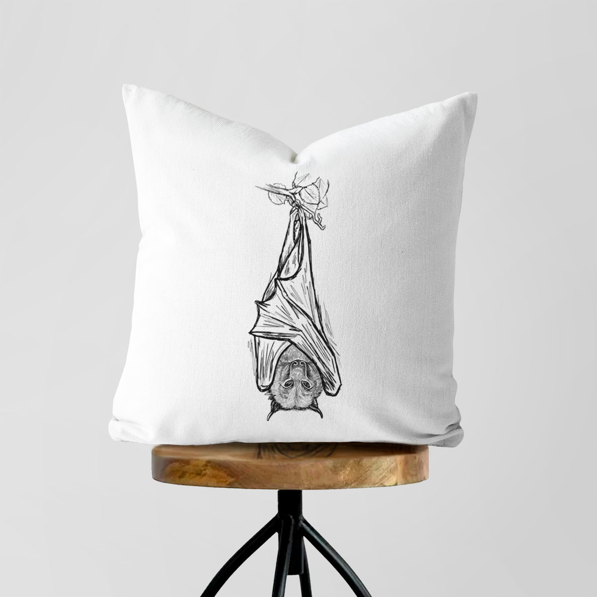 White pillow cover printed with a black ink illustration of a large flying fox bat hanging upside down with its wings wrapped around it, on a wooden stool.