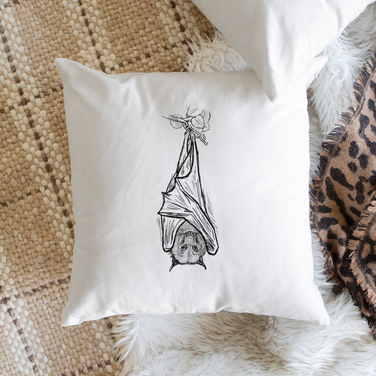 White pillow cover printed with a black ink illustration of a large flying fox bat hanging upside down with its wings wrapped around it, on a woven chair.
