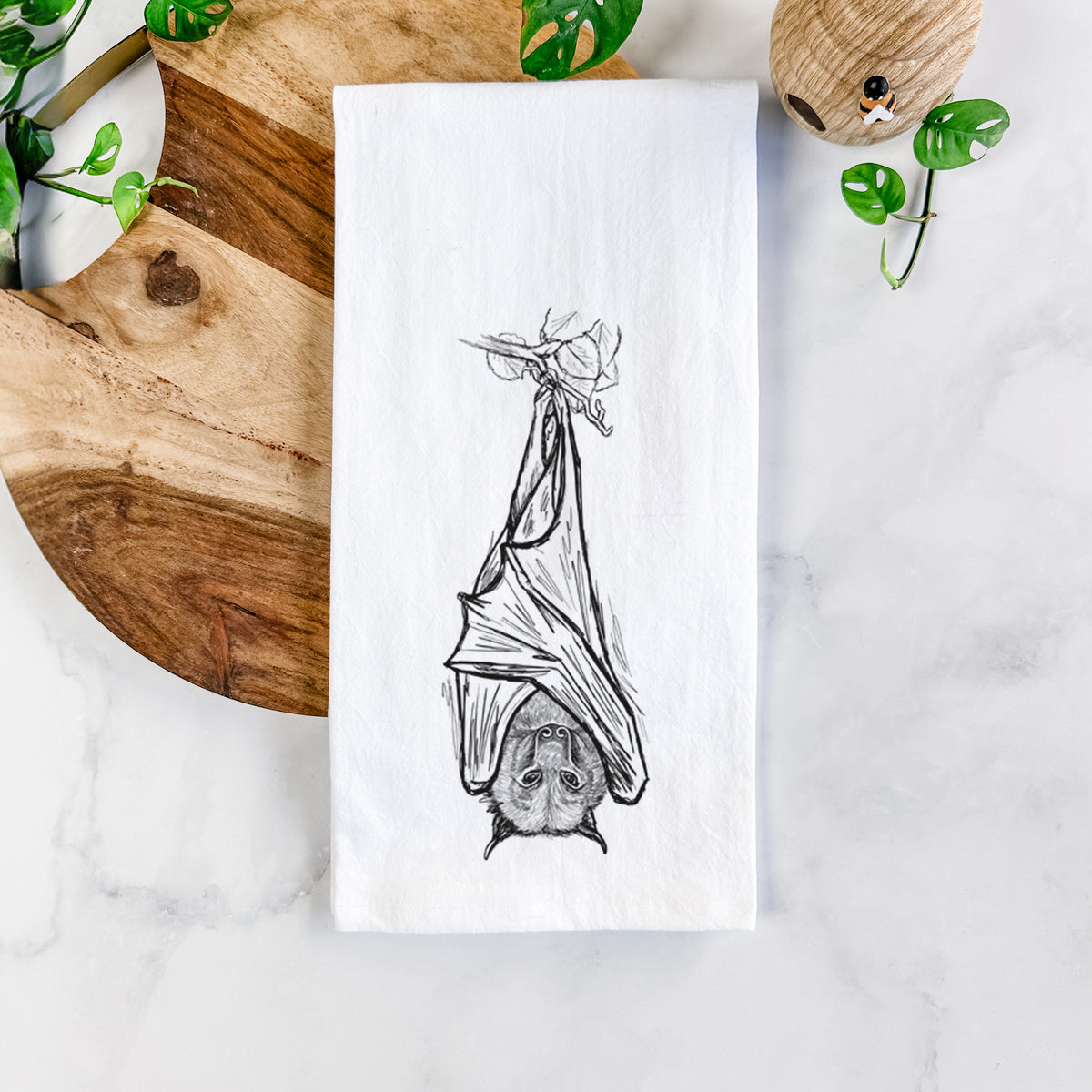 White cotton tea towel printed with a black ink illustration of a large flying fox bat hanging upside down with its wings wrapped around it, on a marble counter beside a round wooden cutting board and greenery.