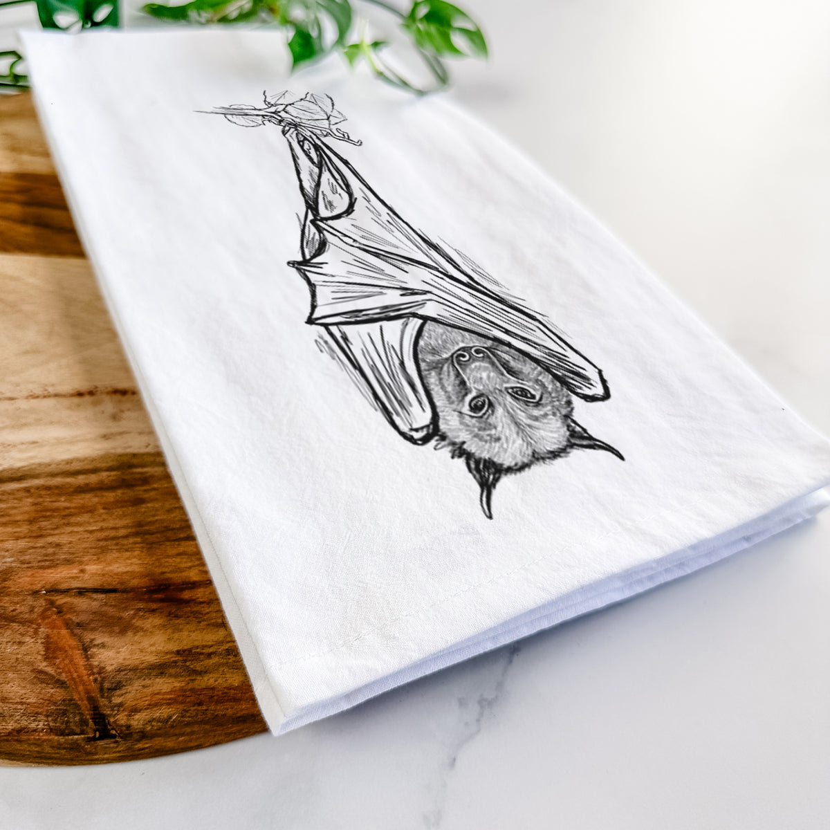 Close-up of a folded white tea towel on a wooden board, showing a black ink illustration of a large flying fox bat hanging upside down with its wings wrapped around it.