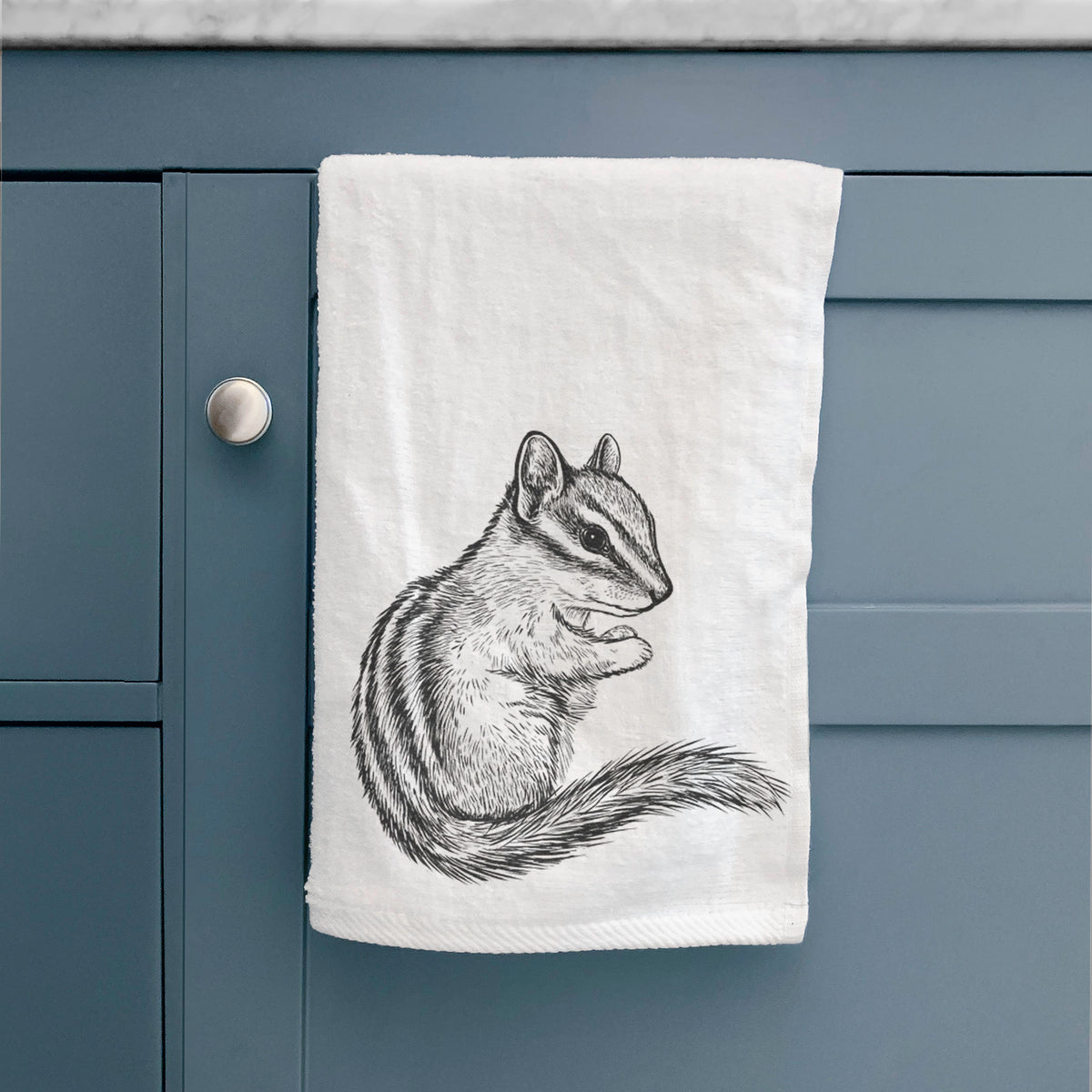 White hand towel printed with a detailed black ink illustration of a least chipmunk sitting and holding food in its paws, hanging on a cabinet door.