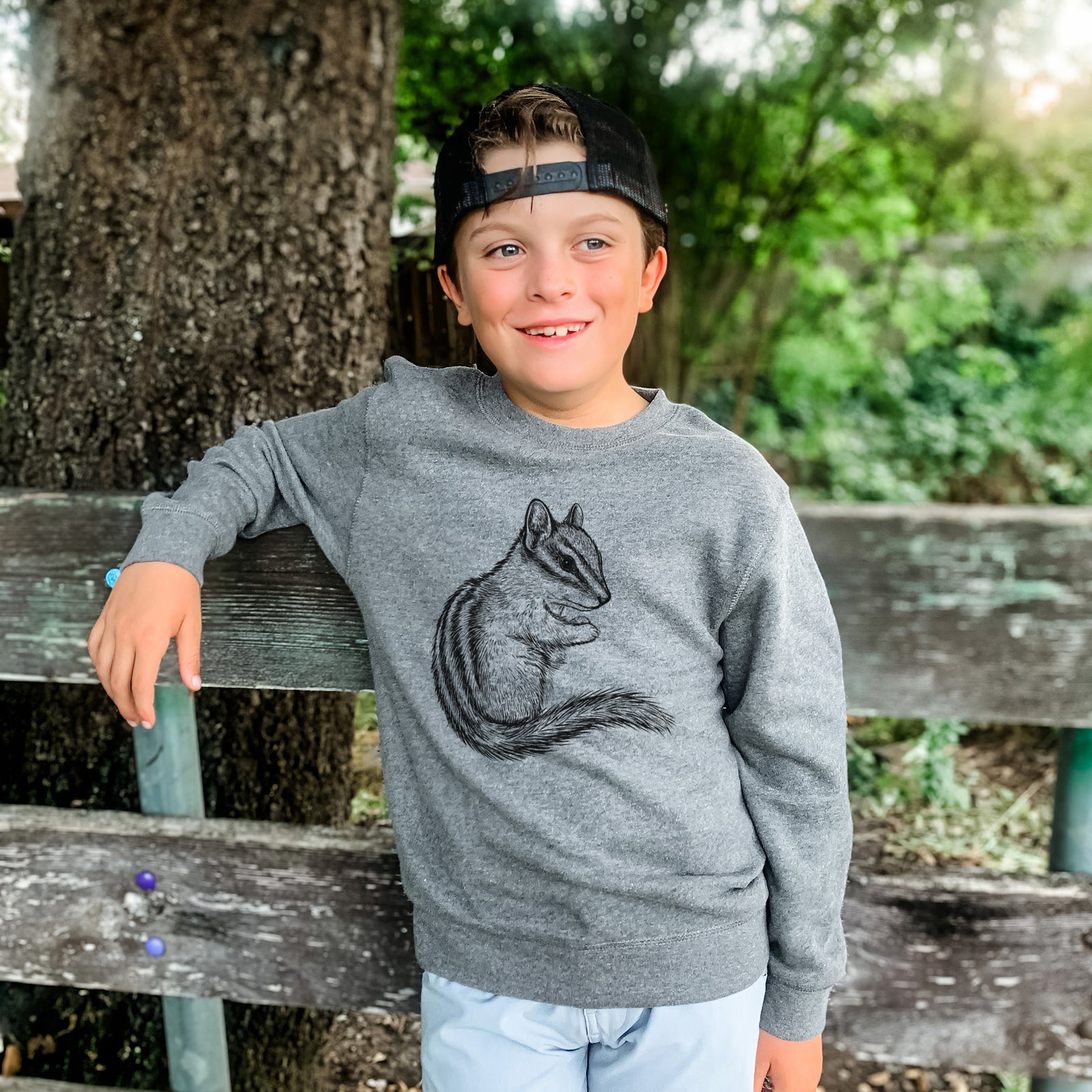 Boy wearing a youth crewneck sweatshirt printed with a detailed black ink illustration of a least chipmunk sitting and holding food in its paws, outdoors.