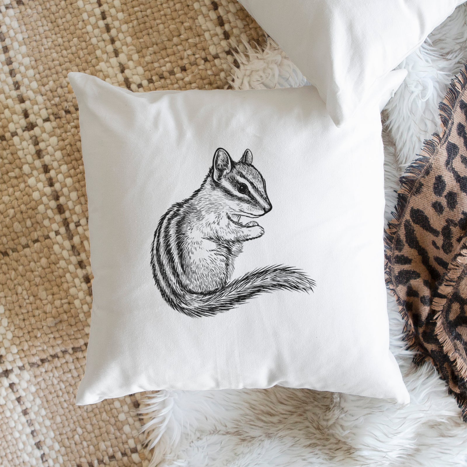 White pillow cover printed with a detailed black ink illustration of a least chipmunk sitting and holding food in its paws, on a bench beside plants.