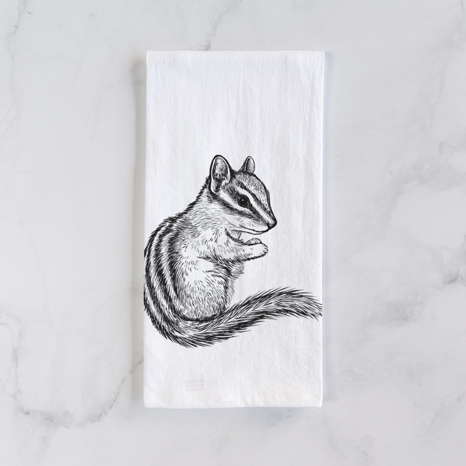 White cotton tea towel printed with a detailed black ink illustration of a least chipmunk sitting and holding food in its paws, on a marble counter beside a round wooden cutting board and greenery.