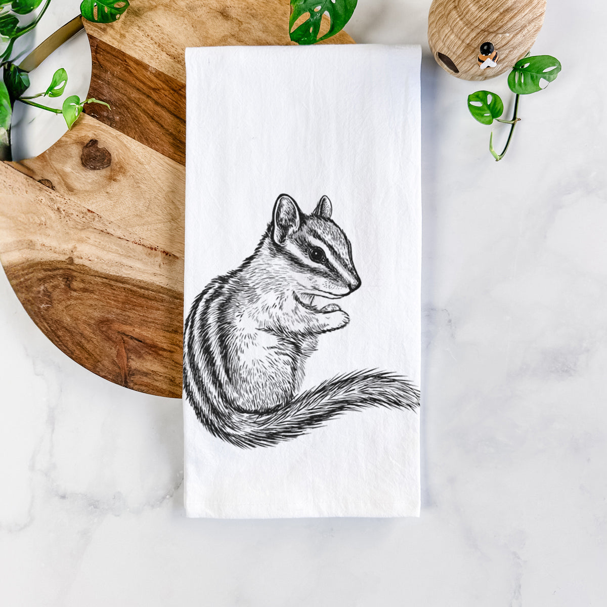White cotton tea towel printed with a detailed black ink illustration of a least chipmunk sitting and holding food in its paws, on a marble counter beside a round wooden cutting board and greenery.