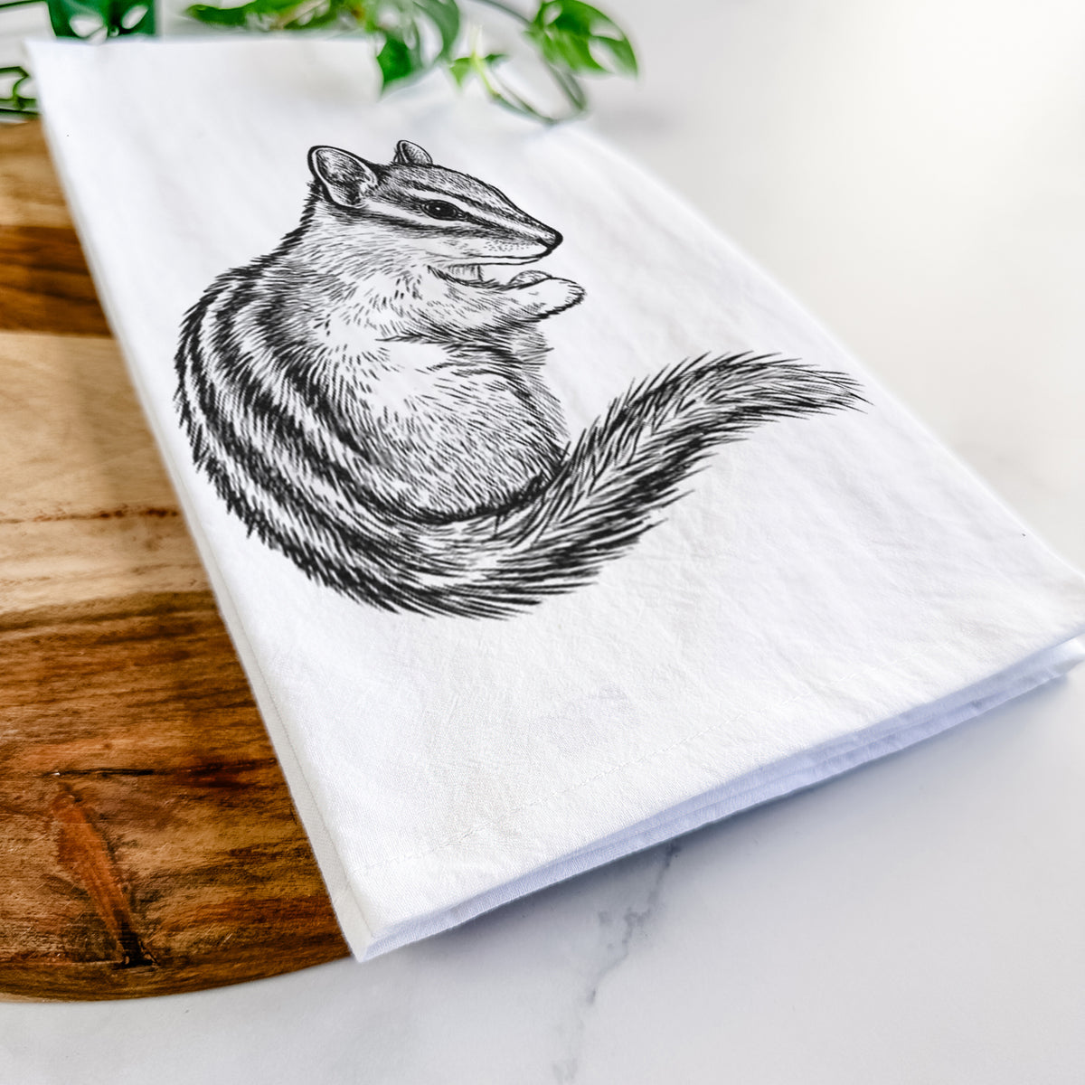 Close-up of a folded white tea towel on a wooden board, showing a detailed black ink illustration of a least chipmunk sitting and holding food in its paws.