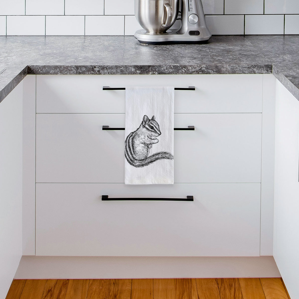 White tea towel printed with a detailed black ink illustration of a least chipmunk sitting and holding food in its paws, hanging from a drawer handle in a bright white kitchen.