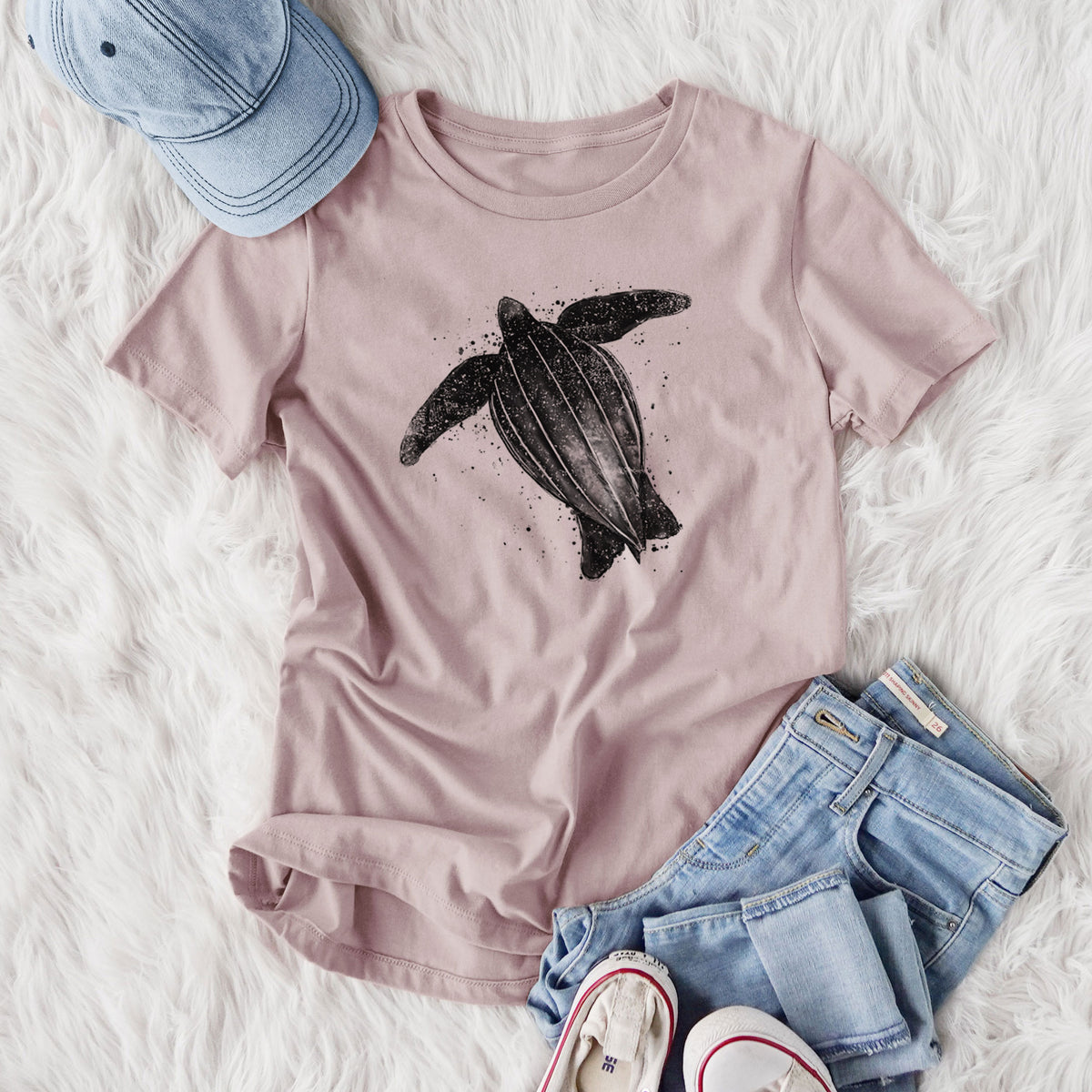 Women's relaxed fit cotton t-shirt printed with a black ink illustration of a leatherback sea turtle swimming, with ink splatters, styled flat on a white faux fur rug with jeans.