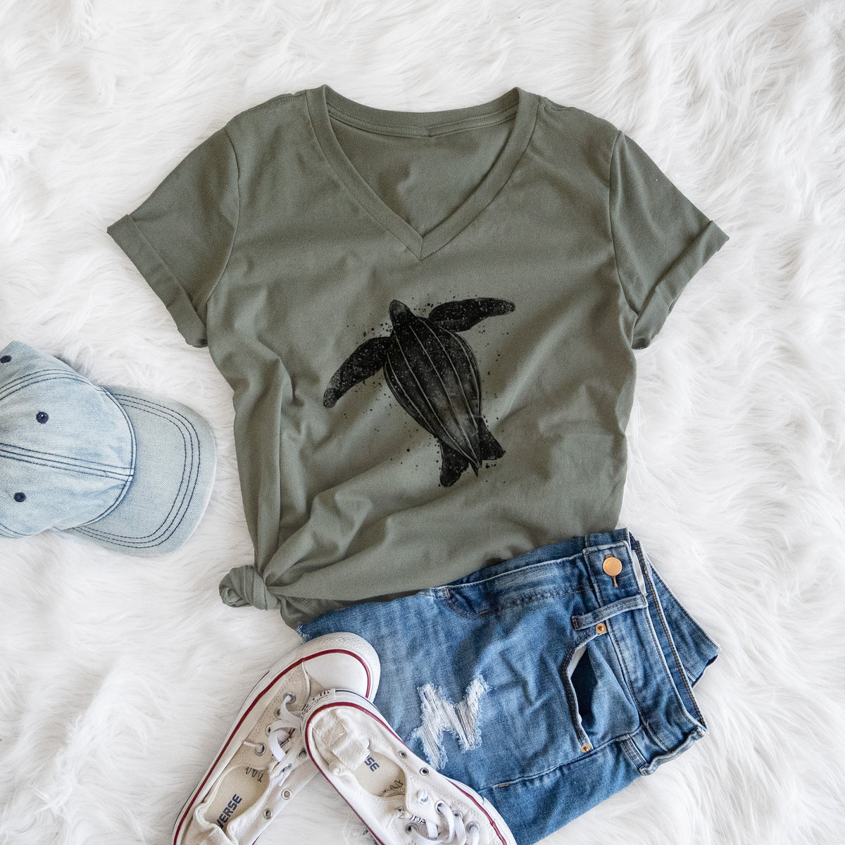 Military green women's relaxed fit cotton V-neck t-shirt printed with a black ink illustration of a leatherback sea turtle swimming, with ink splatters, styled flat on a white faux fur rug with a cap, jeans and sneakers.