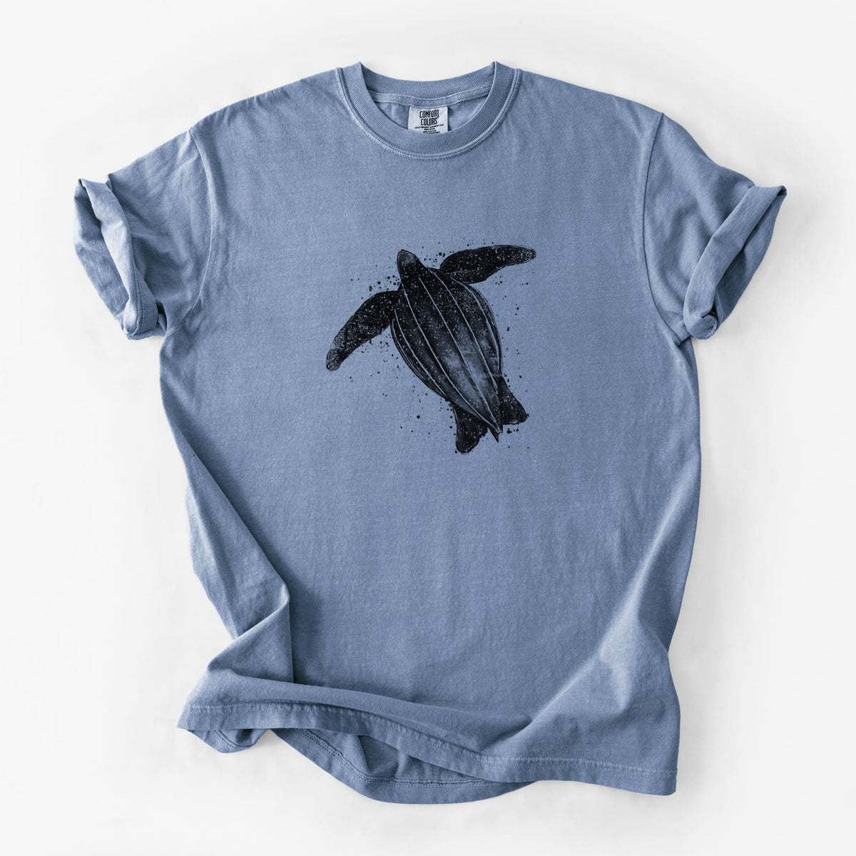 Blue jean Comfort Colors t-shirt laid flat, printed with a black ink illustration of a leatherback sea turtle swimming, with ink splatters.