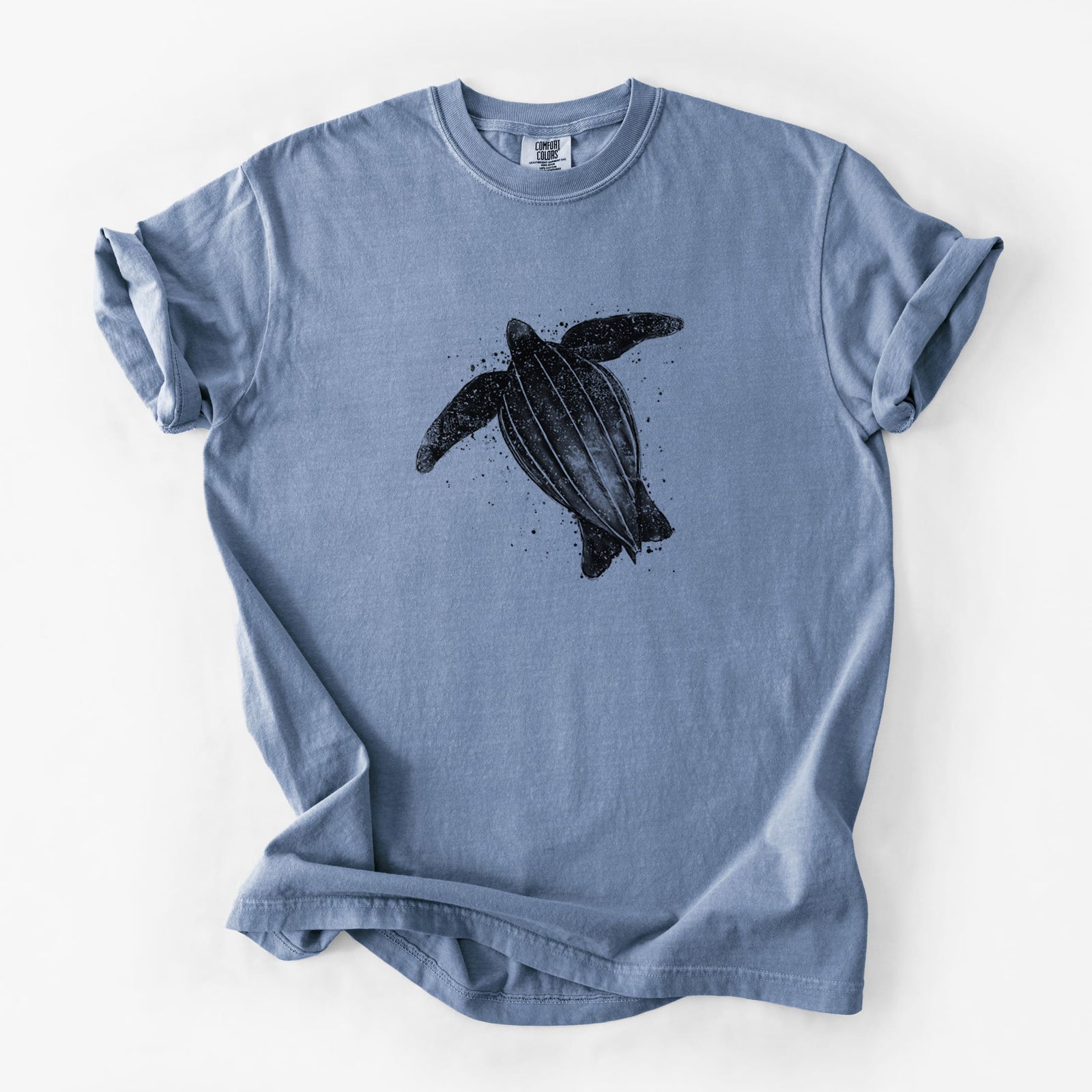 Blue jean Comfort Colors t-shirt laid flat, printed with a black ink illustration of a leatherback sea turtle swimming, with ink splatters.