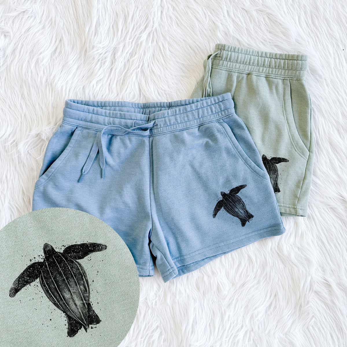 Two pairs of Cali Wave shorts in different colors printed with a black ink illustration of a leatherback sea turtle swimming, with ink splatters, with a close-up of the print.