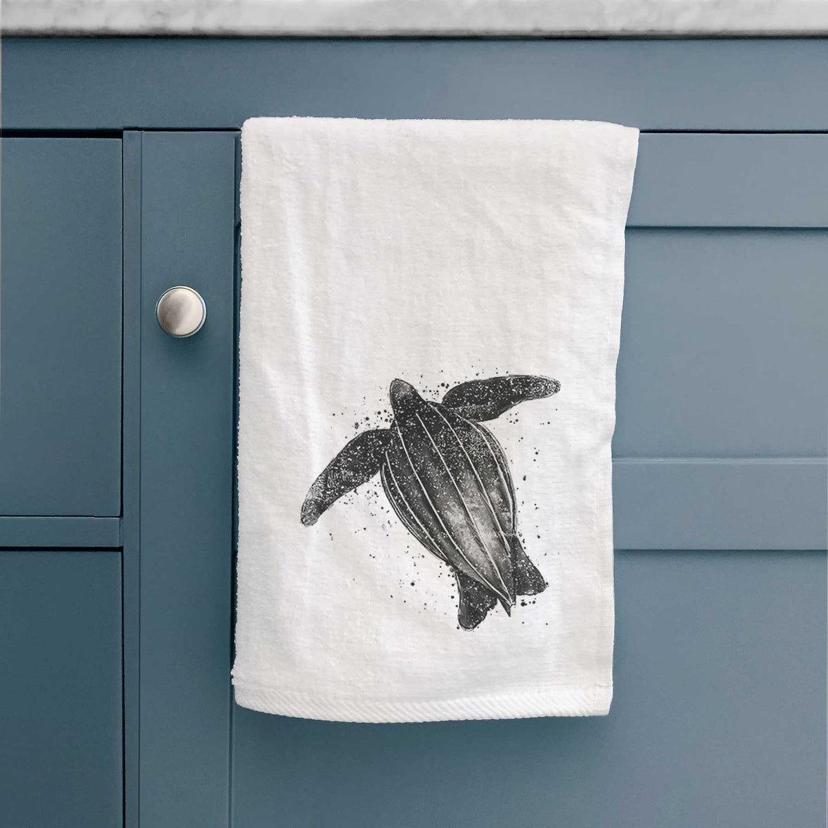 White hand towel printed with a black ink illustration of a leatherback sea turtle swimming, with ink splatters, hanging on a cabinet door.
