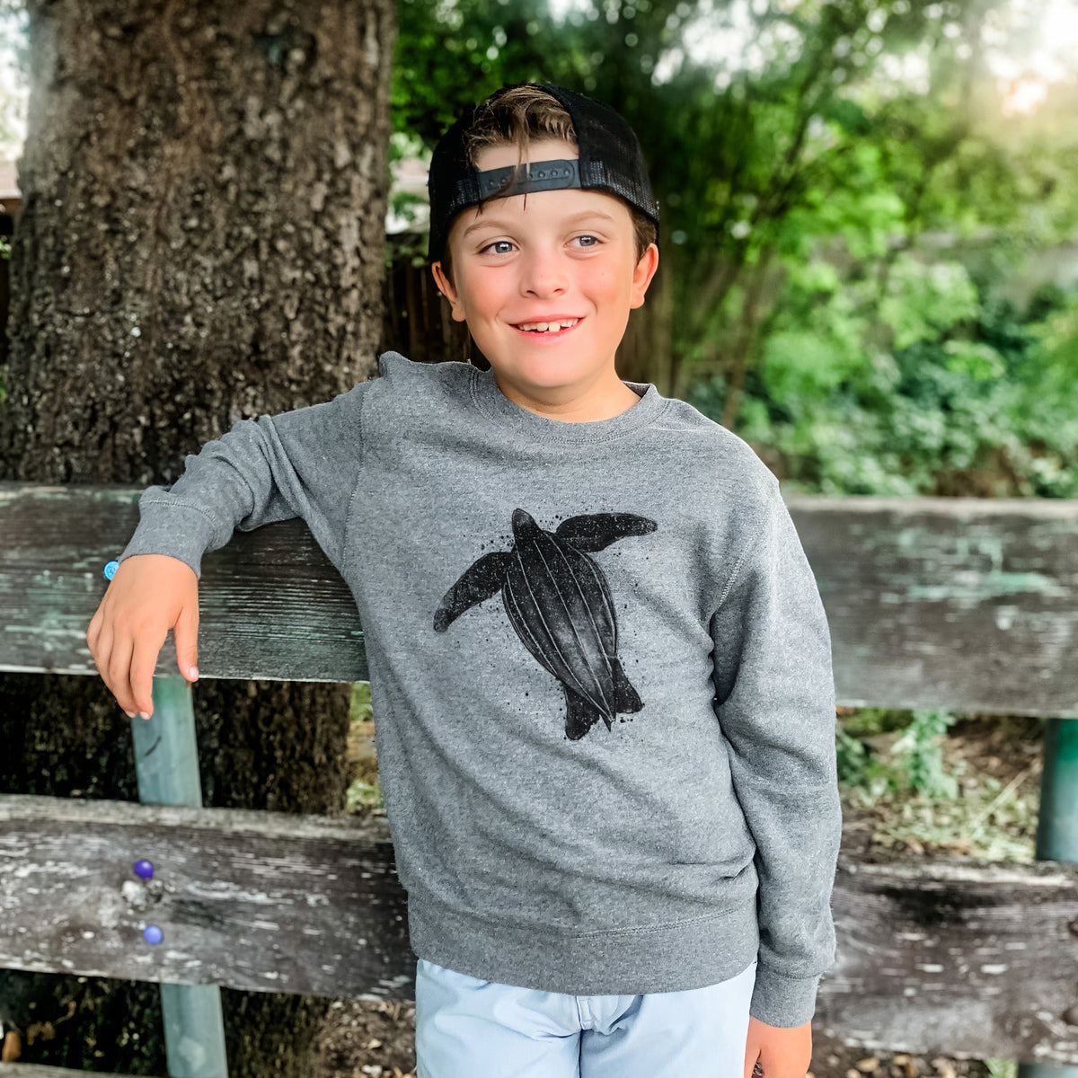 Boy wearing a youth crewneck sweatshirt printed with a black ink illustration of a leatherback sea turtle swimming, with ink splatters, outdoors.