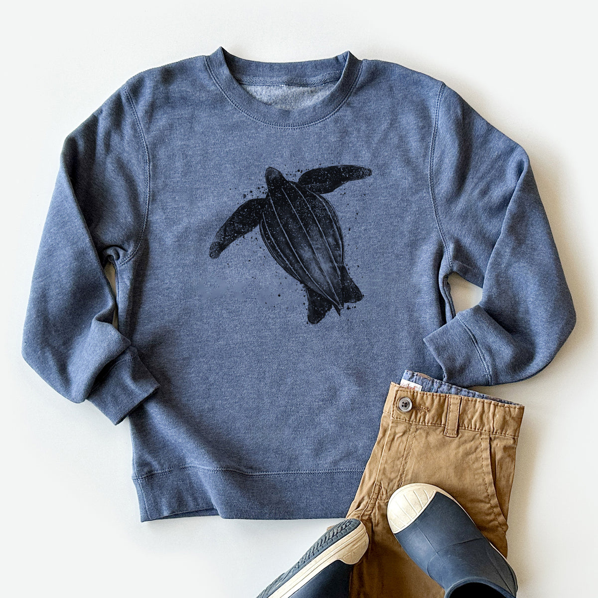 Youth crewneck sweatshirt printed with a black ink illustration of a leatherback sea turtle swimming, with ink splatters, styled flat with khaki pants and shoes.