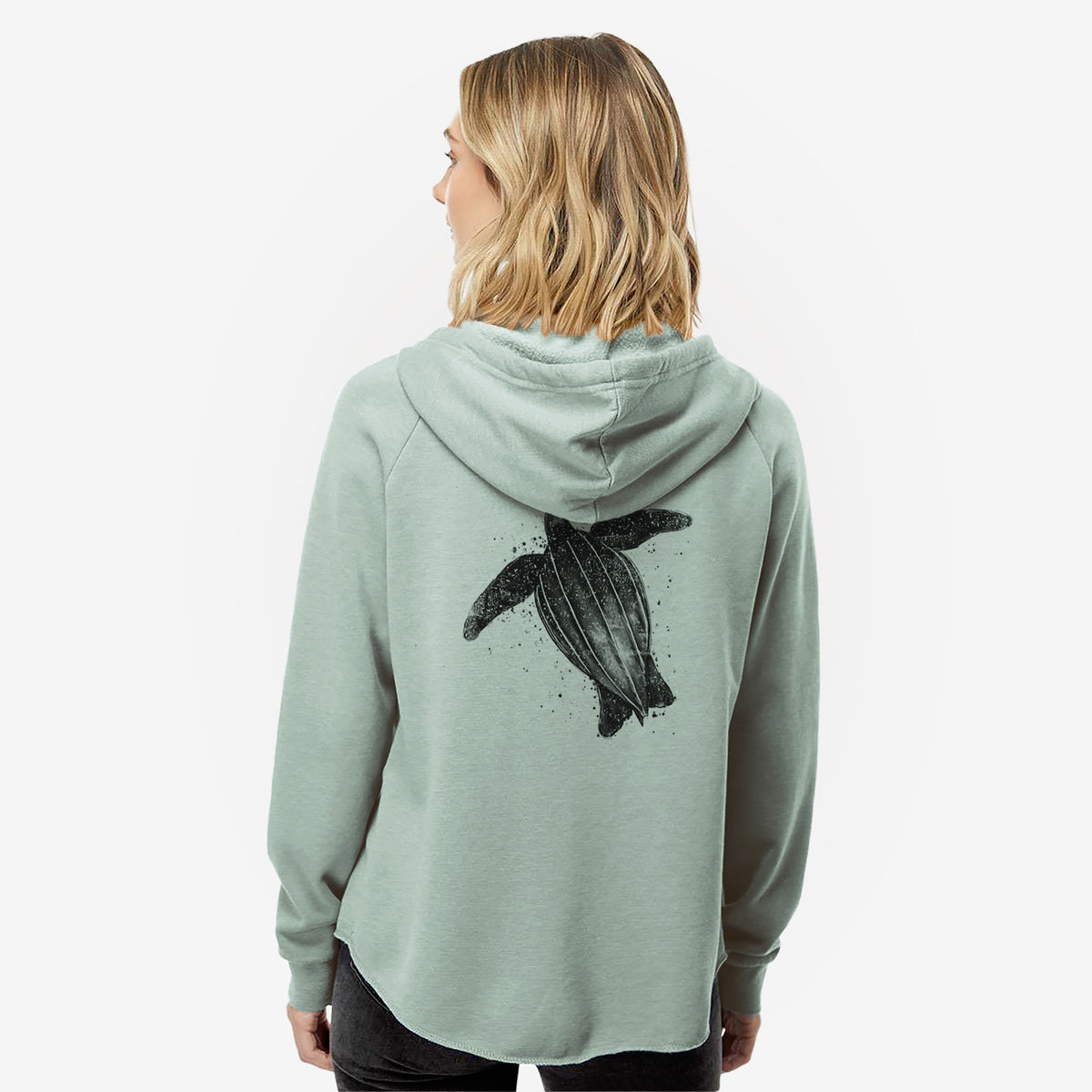 Woman wearing a sage green women's Cali Wave zip-up sweatshirt printed with a black ink illustration of a leatherback sea turtle swimming, with ink splatters, seen from behind.