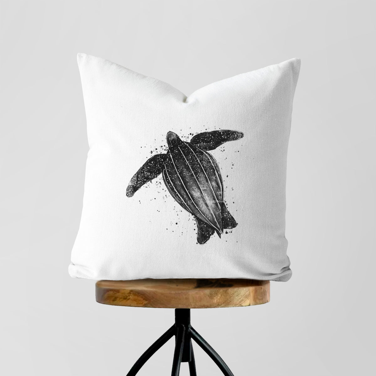 White pillow cover printed with a black ink illustration of a leatherback sea turtle swimming, with ink splatters, on a wooden stool.