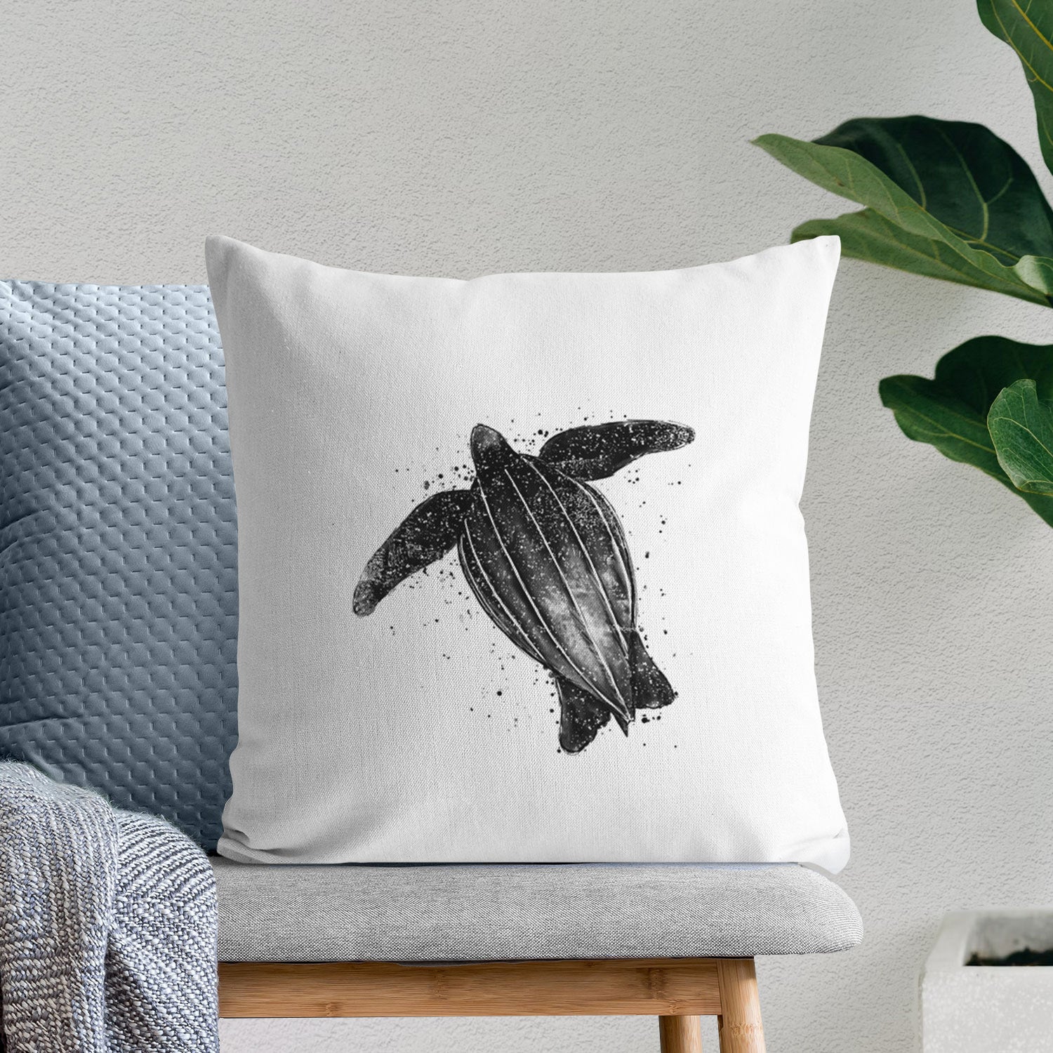 White pillow cover printed with a black ink illustration of a leatherback sea turtle swimming, with ink splatters, on a bench beside plants.