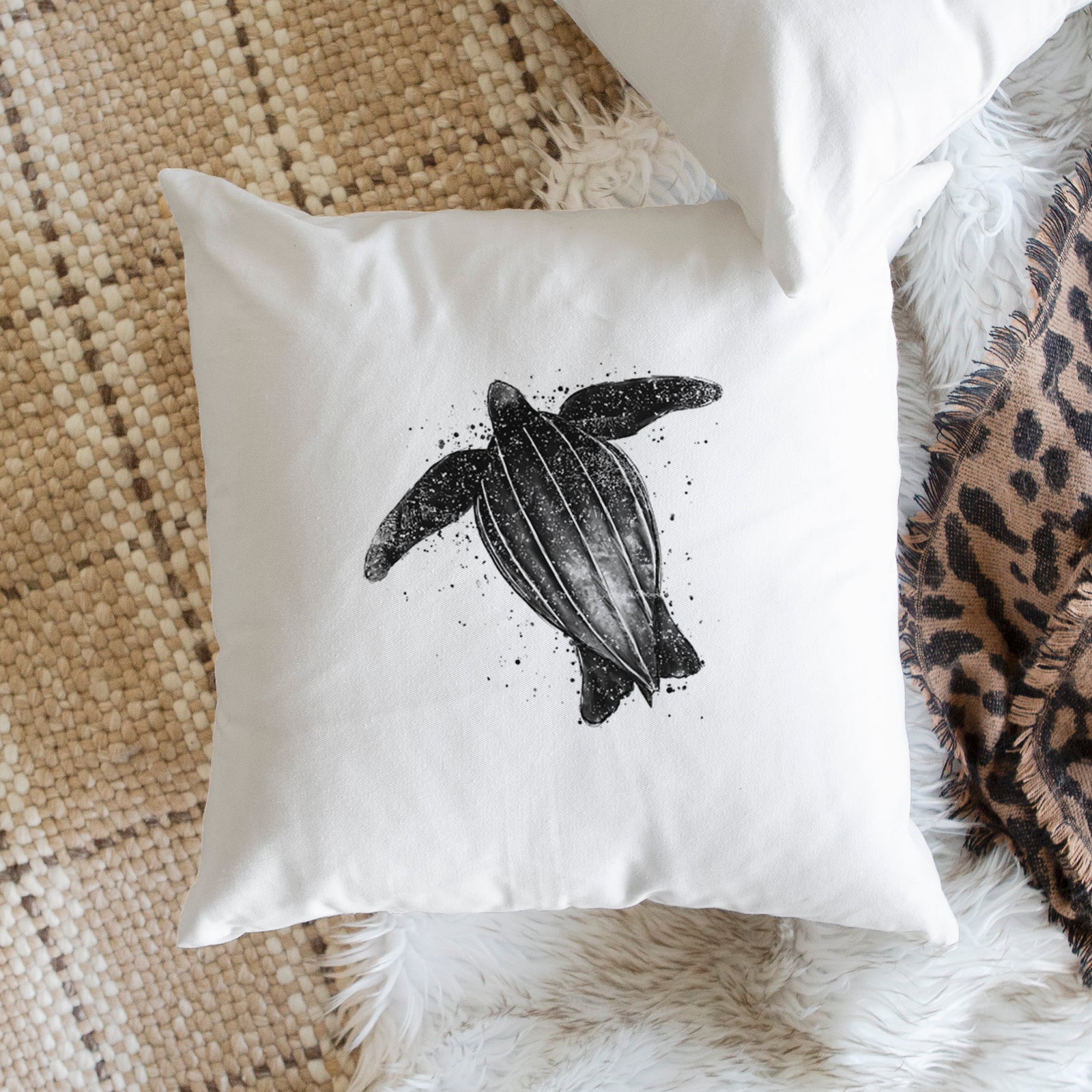 White pillow cover printed with a black ink illustration of a leatherback sea turtle swimming, with ink splatters, on a bench beside plants.