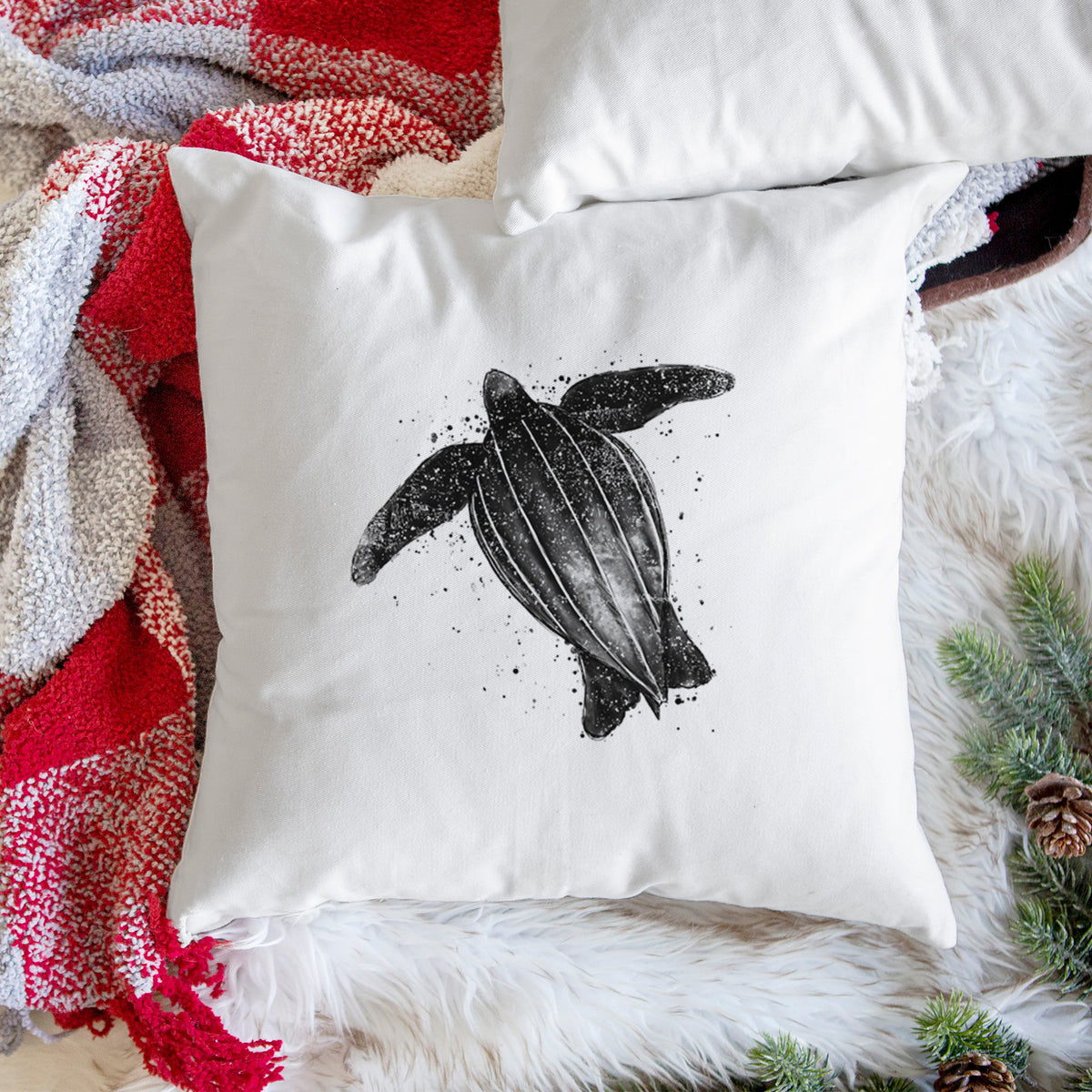 White pillow cover printed with a black ink illustration of a leatherback sea turtle swimming, with ink splatters, on a woven chair.