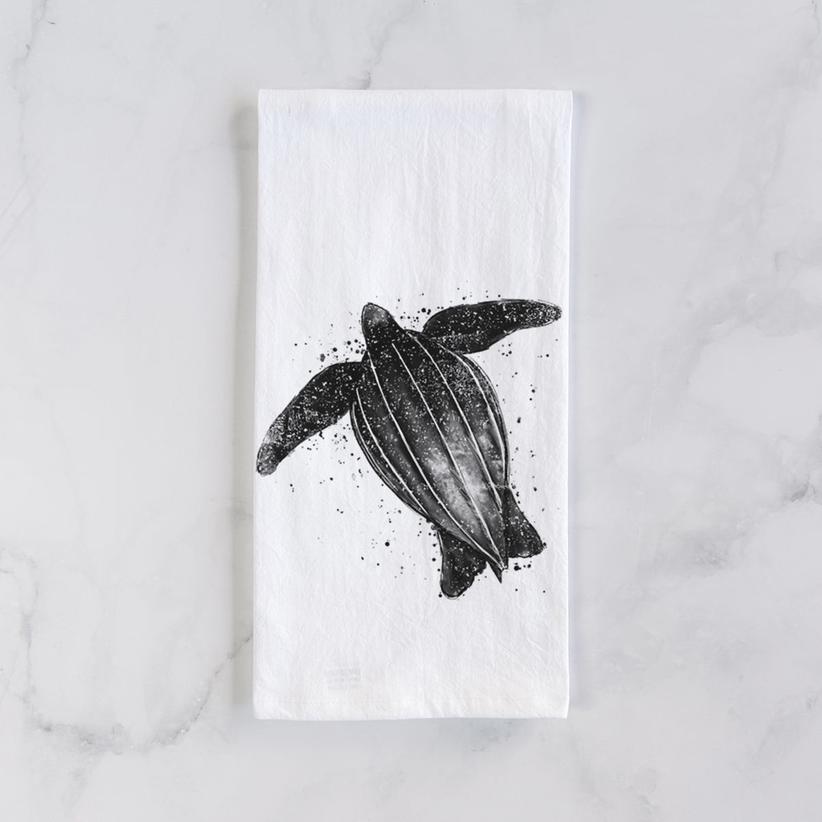 White cotton tea towel laid flat on marble, printed with a black ink illustration of a leatherback sea turtle swimming, with ink splatters.