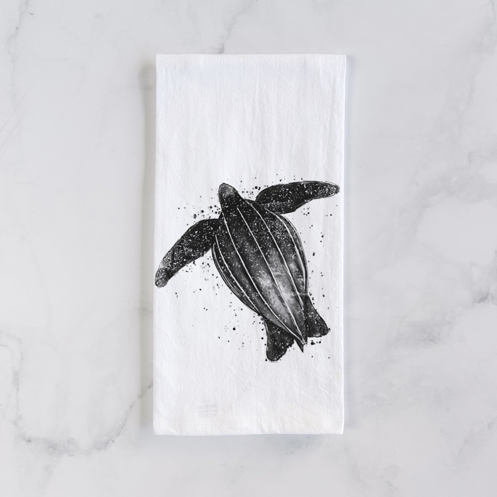 White cotton tea towel printed with a black ink illustration of a leatherback sea turtle swimming, with ink splatters, on a marble counter beside a round wooden cutting board and greenery.